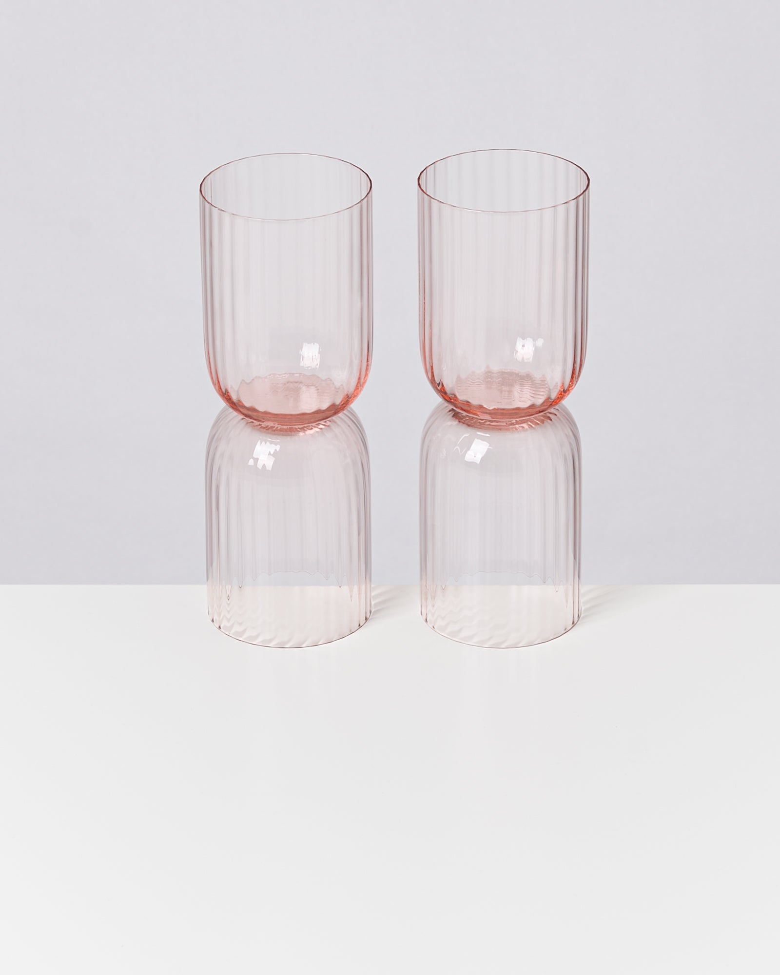 Zwei blassrosa, gerillte Gläser stehen auf einem weißen Tisch vor einem grauen Hintergrund.