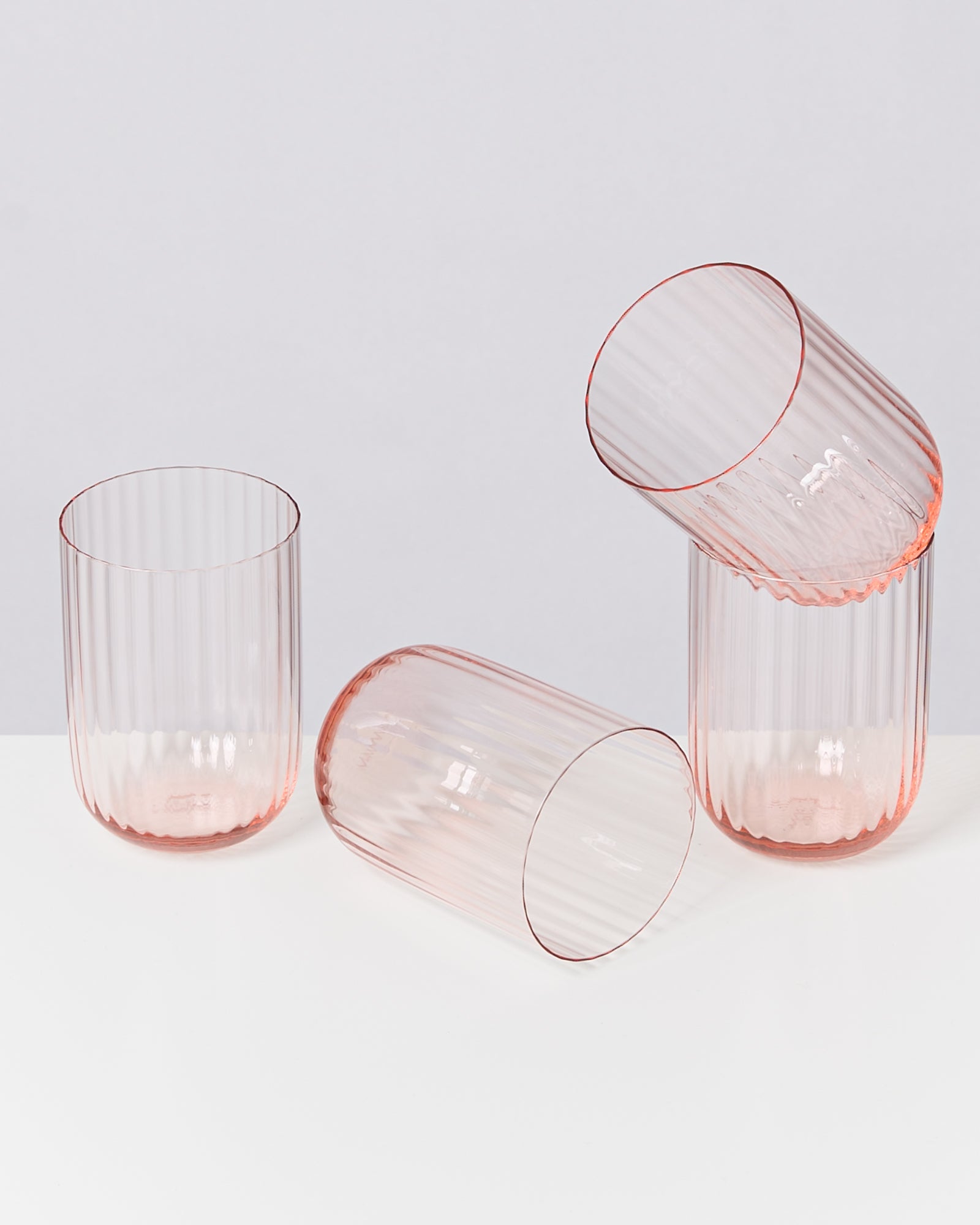 Vier rosafarbene, gerippte Gläser, drei stehen, eines liegt auf einem weißen Untergrund.