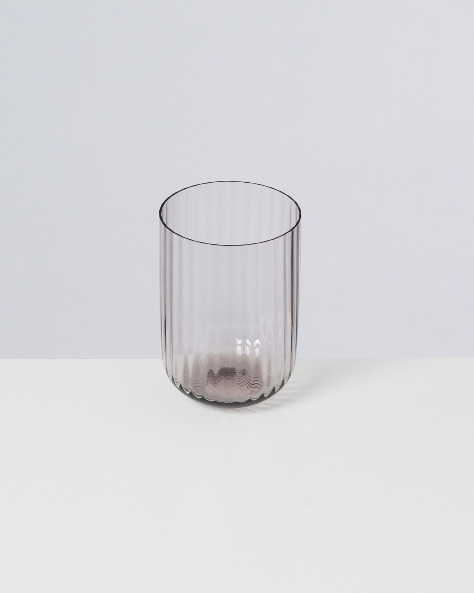 Ein durchsichtiges, geripptes Trinkglas steht auf einem weißen Tisch vor einem grauen Hintergrund.