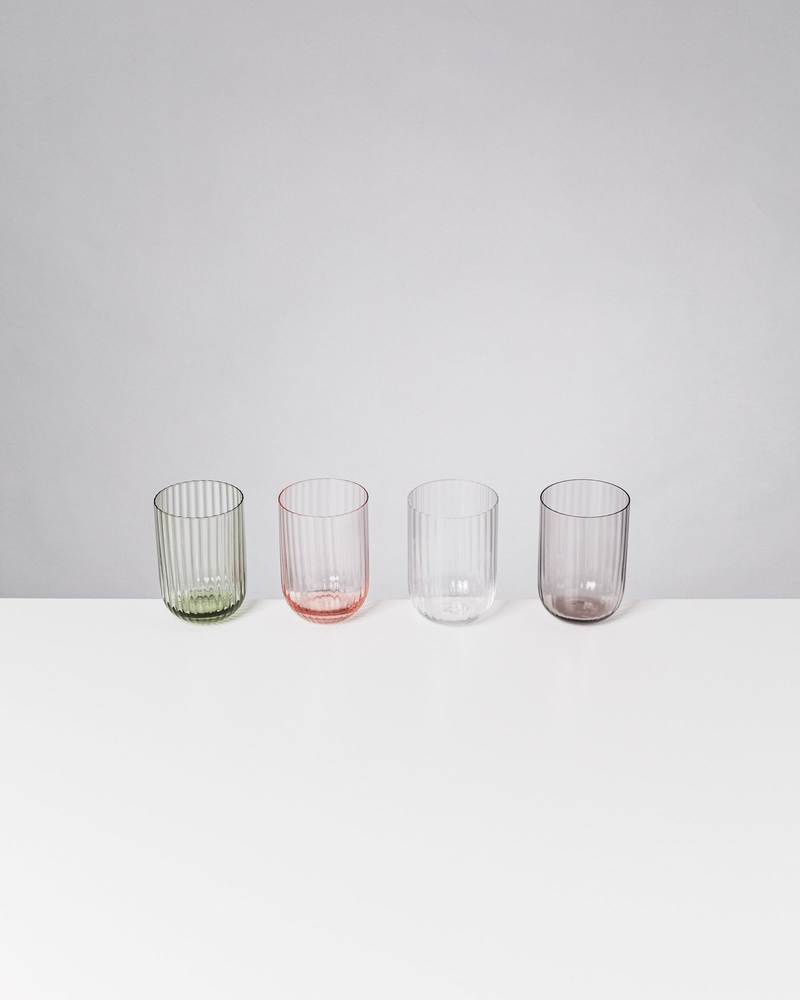 Vier geriffelte Gläser in verschiedenen Farben stehen in einer Linie auf einem weißen Tisch.