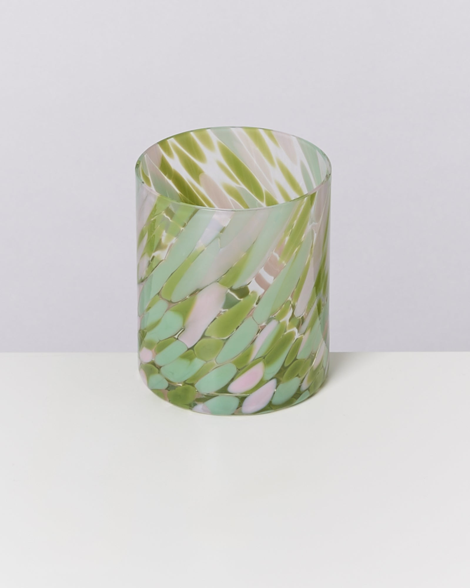 Ein zylindrisches Glasgefäß mit grünem, rosa und weißem Muster steht auf einem weißen Tisch vor grauem Hintergrund.