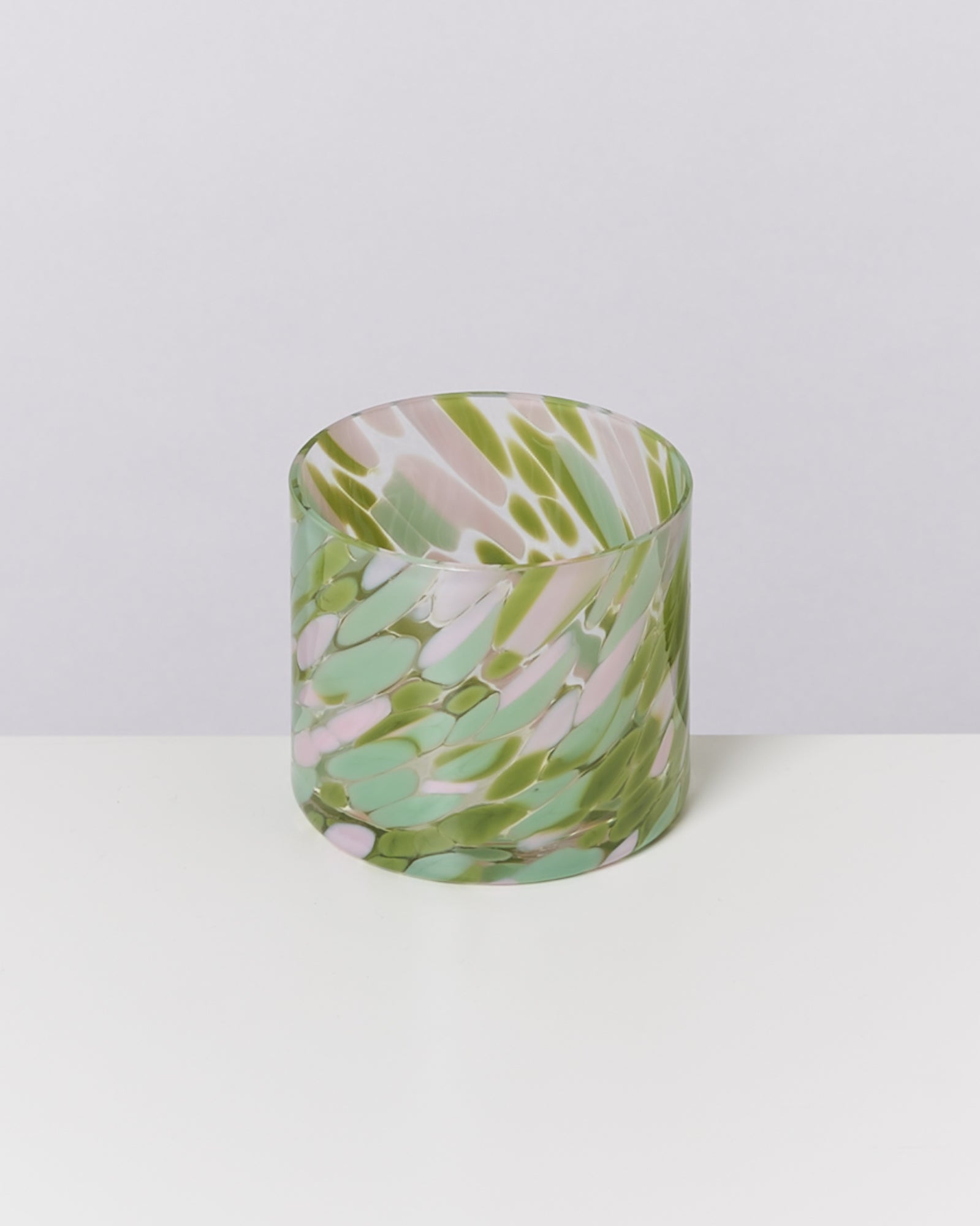 Zylinderförmiges Glas mit grünem, rosa und weißem Muster steht auf einem weißen Untergrund.