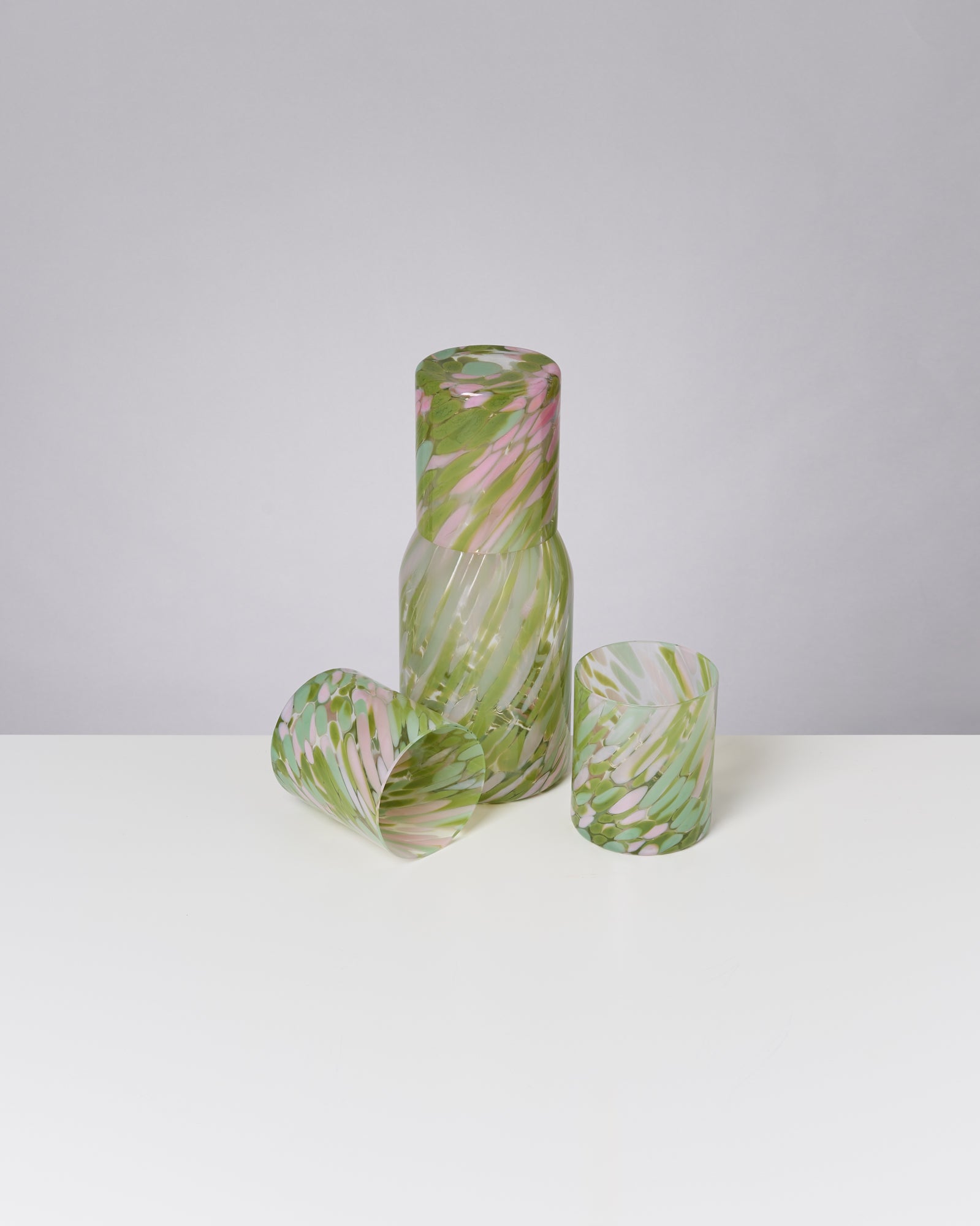 Eine gläserne Karaffe und zwei Gläser mit grün-rosa Muster stehen auf einem weißen Tisch.