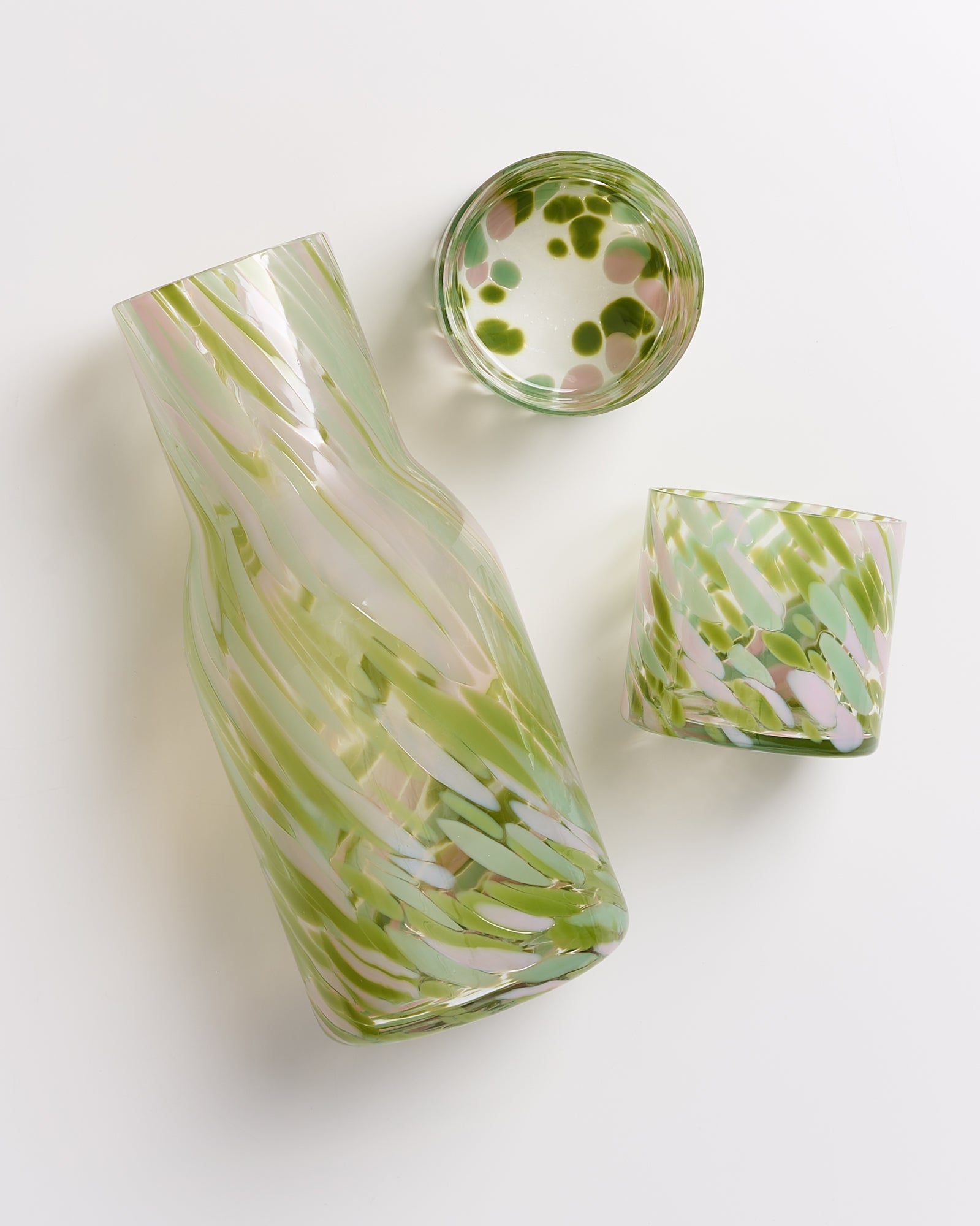 Glasflasche und zwei Trinkgläser mit grünem und rosa Muster auf weißem Hintergrund, von oben fotografiert.