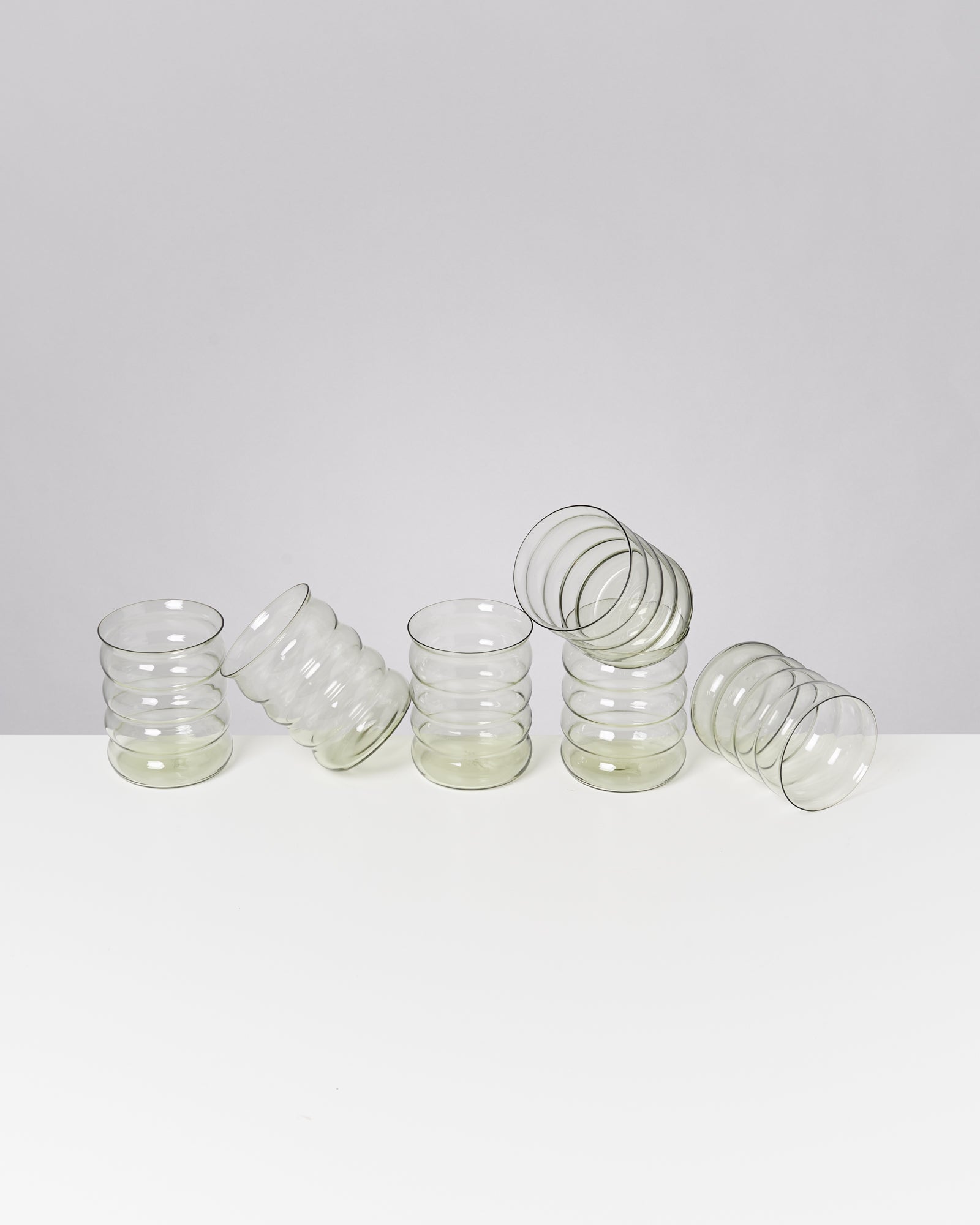 Fünf durchsichtige, gewellte Gläser stehen und liegen auf einem weißen Tisch vor grauem Hintergrund.