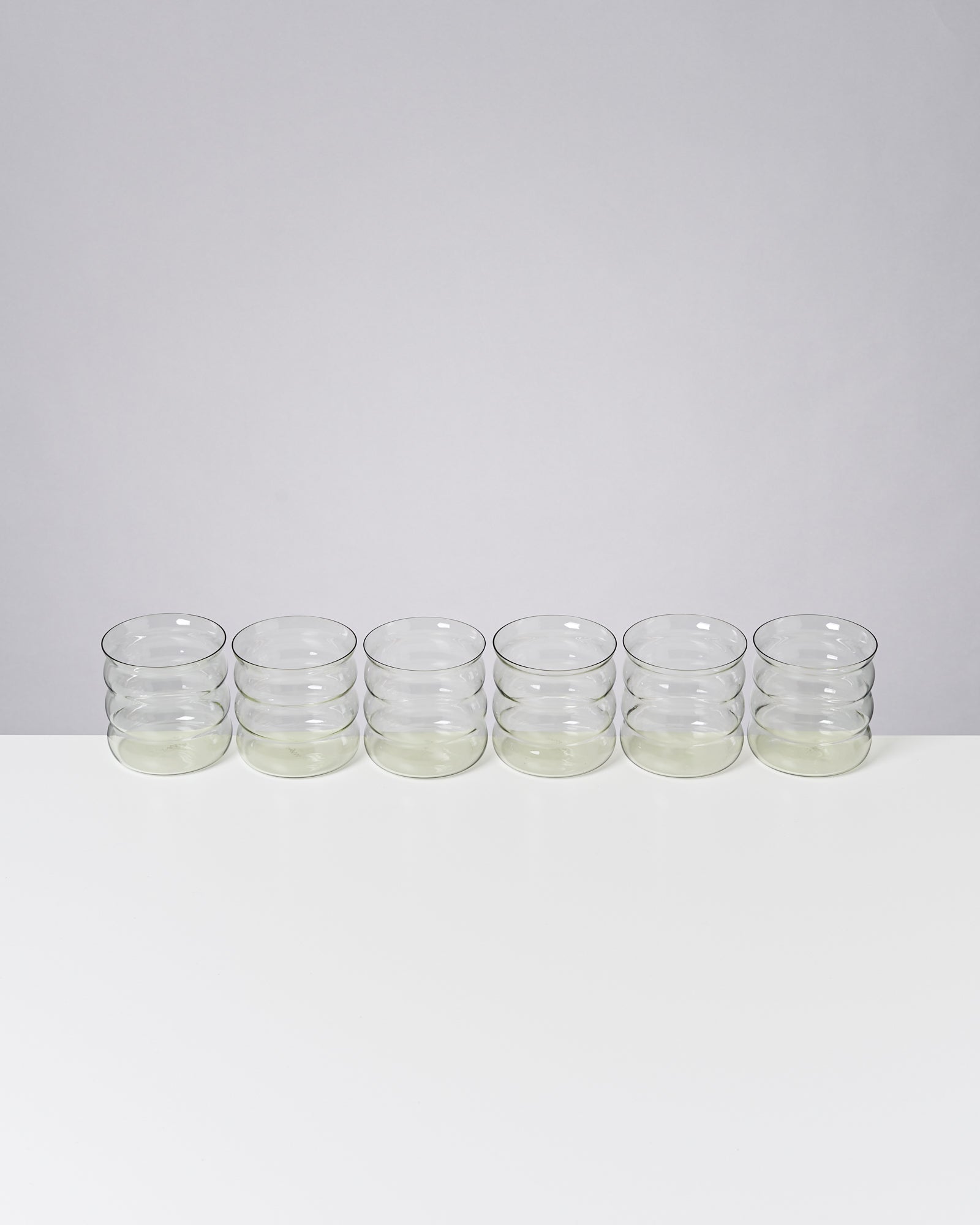 Sechs leere, stapelbare Gläser stehen in einer Reihe auf einem weißen Tisch vor grauem Hintergrund.