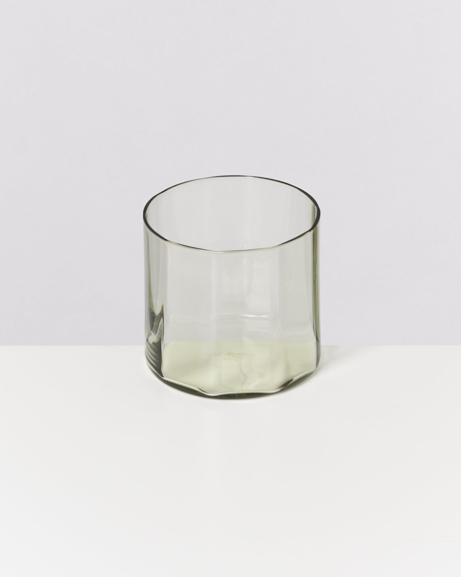 Transparenter, zylindrischer Glasbecher auf einer weißen Oberfläche vor neutralem Hintergrund.