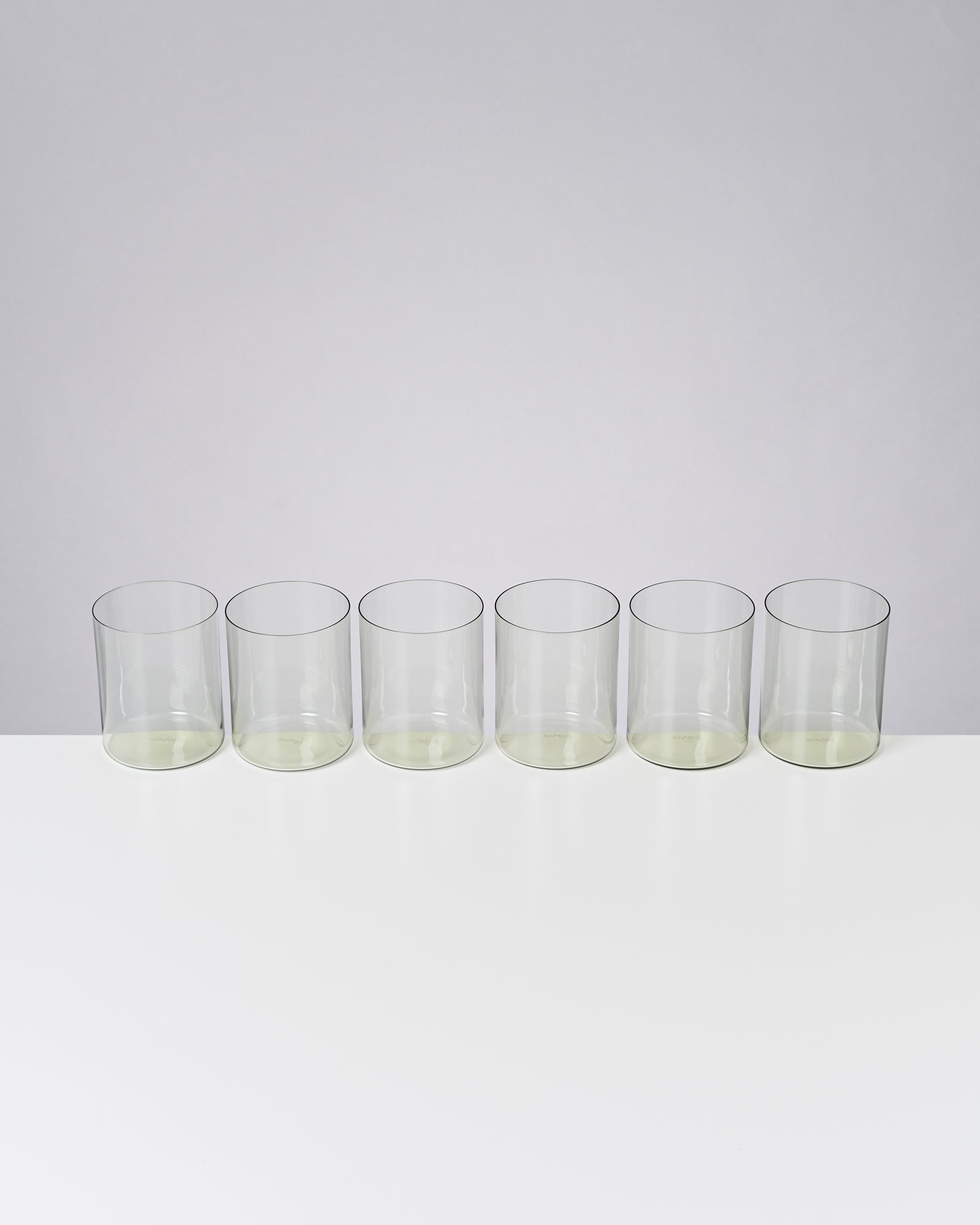 Sechs leere, transparente Gläser stehen in einer Reihe auf einer weißen Oberfläche vor grauem Hintergrund.