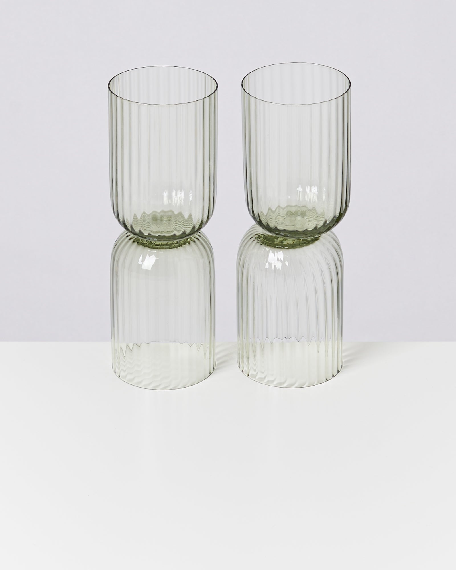 Zwei grüne, geriffelte Trinkgläser stehen auf einem weißen Tisch mit ihrer Reflektion darunter.
