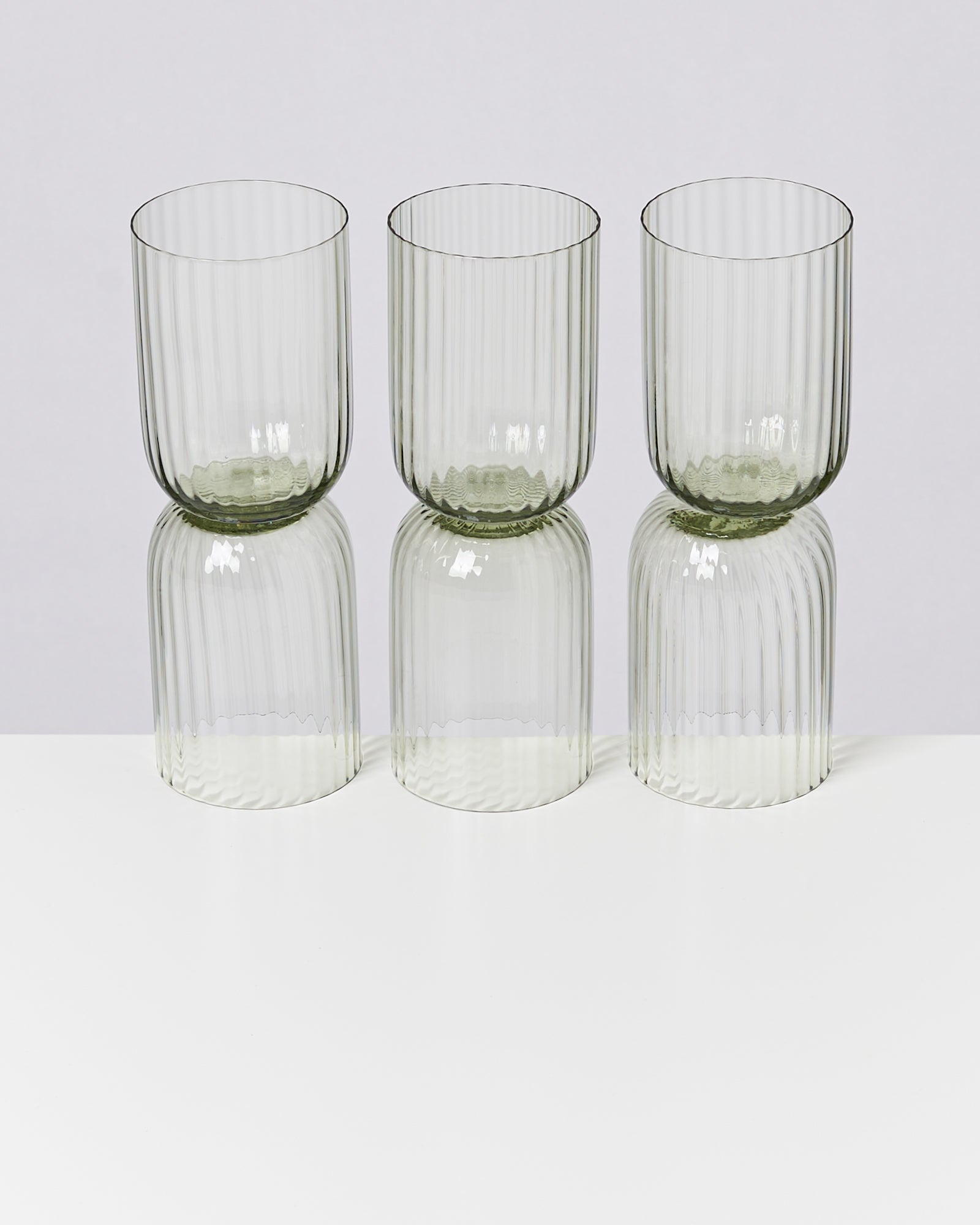 Drei geriffelte, durchsichtige Trinkgläser stehen in einer Reihe auf einem weißen Tisch vor einem grauen Hintergrund.