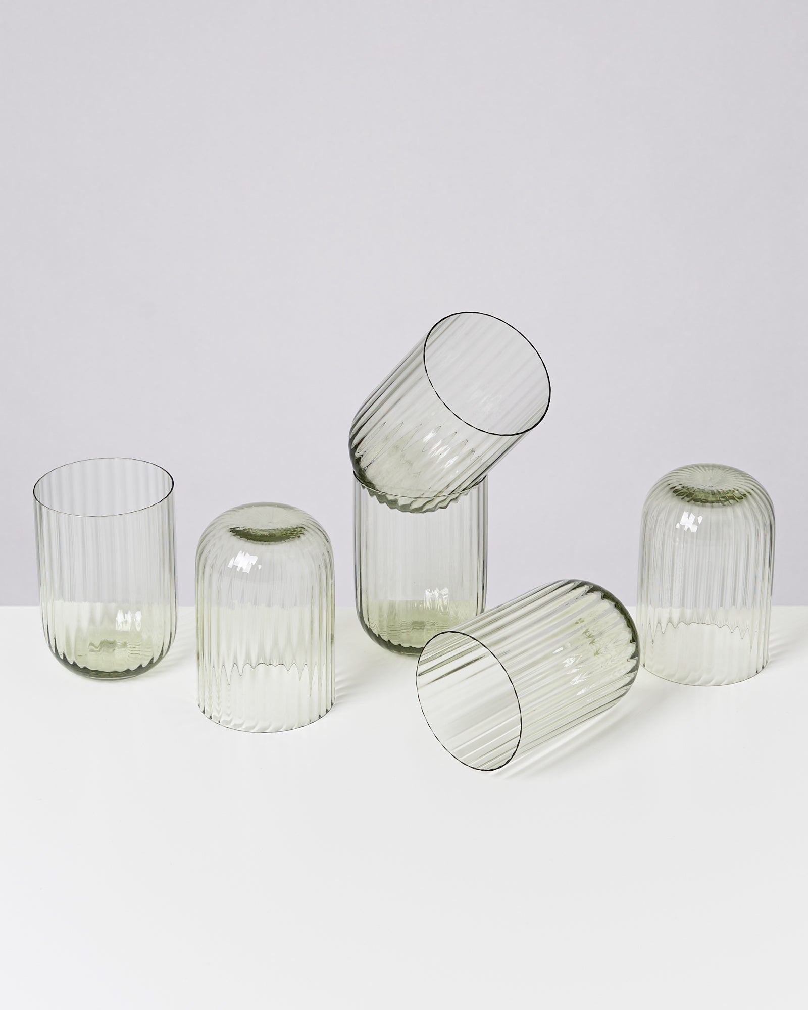 Fünf transparente, geriffelte Gläser stehen und liegen schief auf einer weißen Oberfläche.