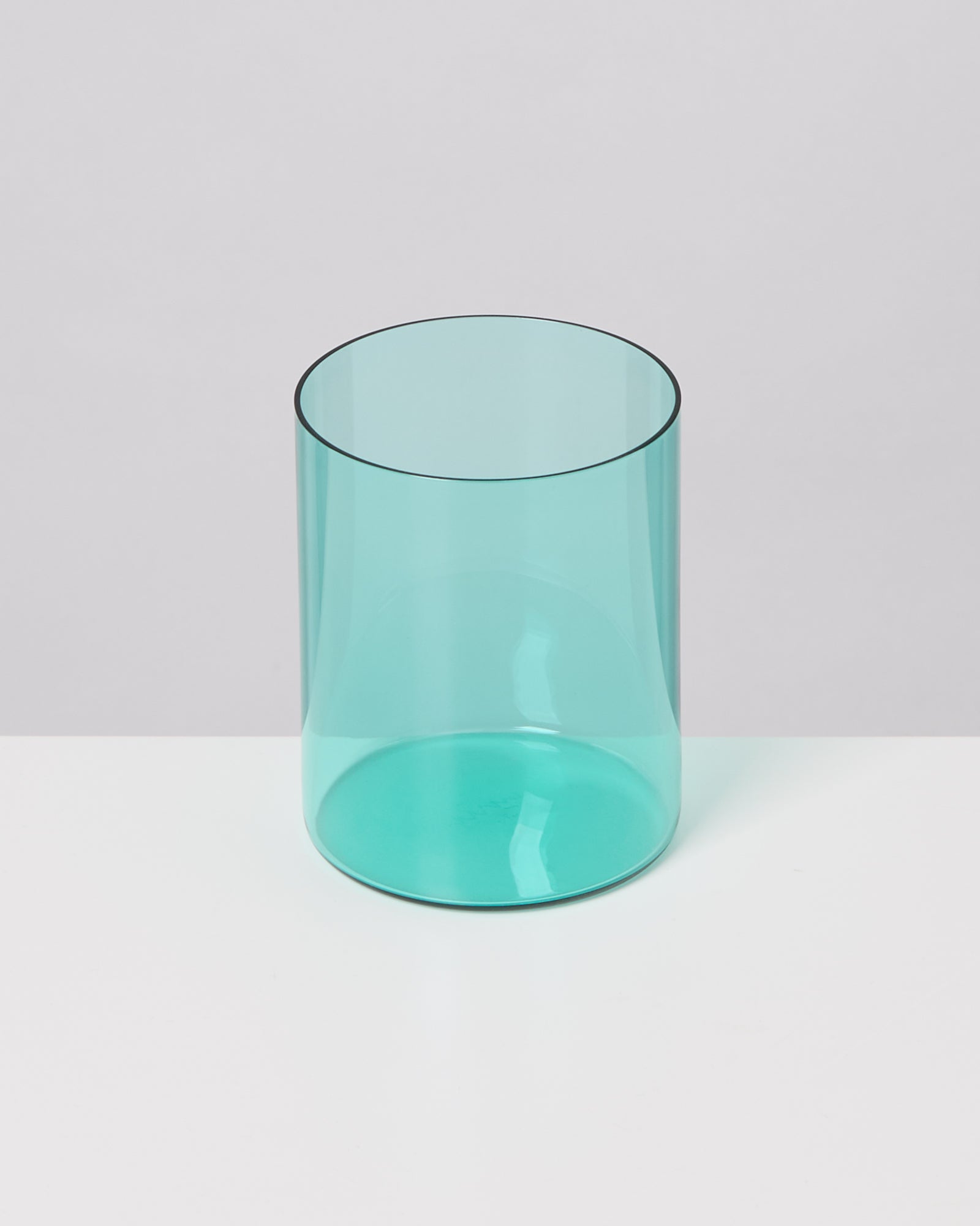 Ein zylindrisches, transparentes, türkisfarbenes Glas steht auf einer weißen Oberfläche vor einem grauen Hintergrund.