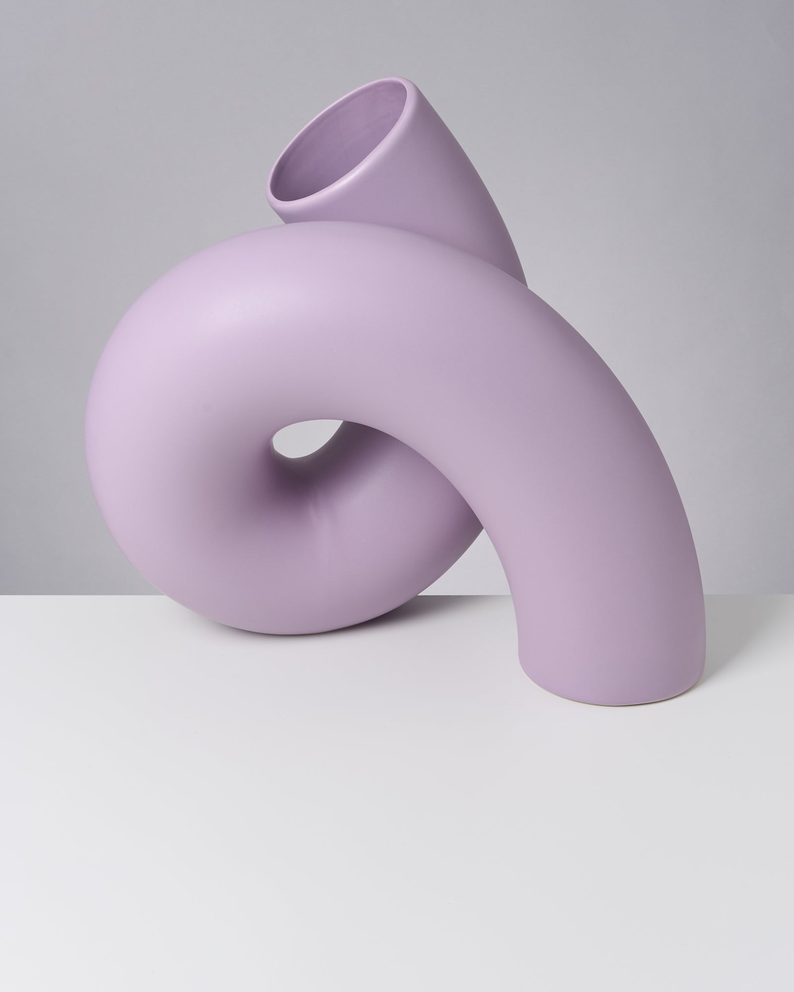 Lila, abstrakt geformte Vase auf weißem Tisch vor grauem Hintergrund.