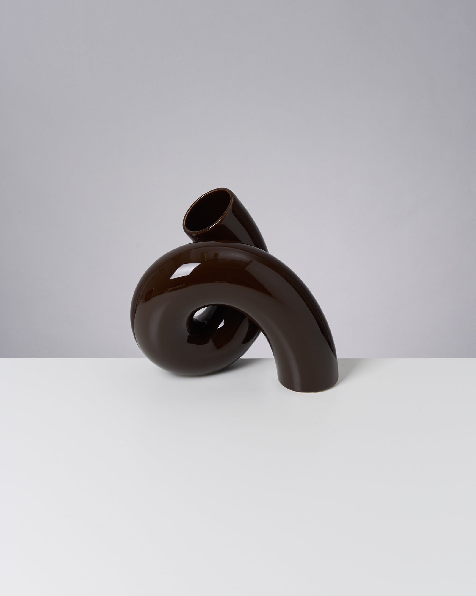 Braune, gebogene Keramikvase mit glänzender Oberfläche auf einem weißen Tisch vor grauem Hintergrund.