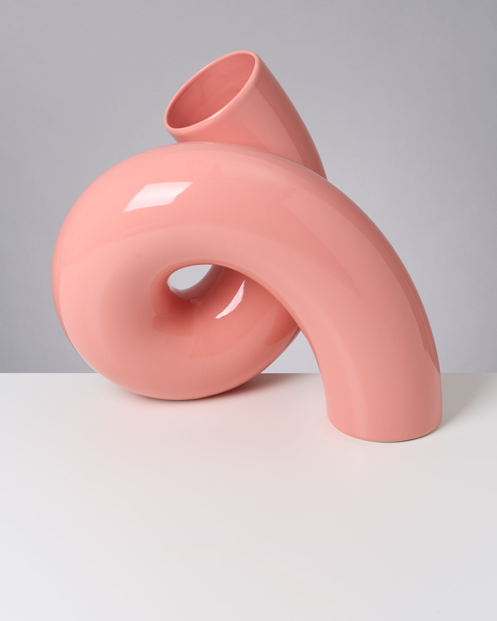 Rosafarbene, moderne Keramikvase mit geschwungenem Design auf einem weißen Tisch vor grauem Hintergrund.