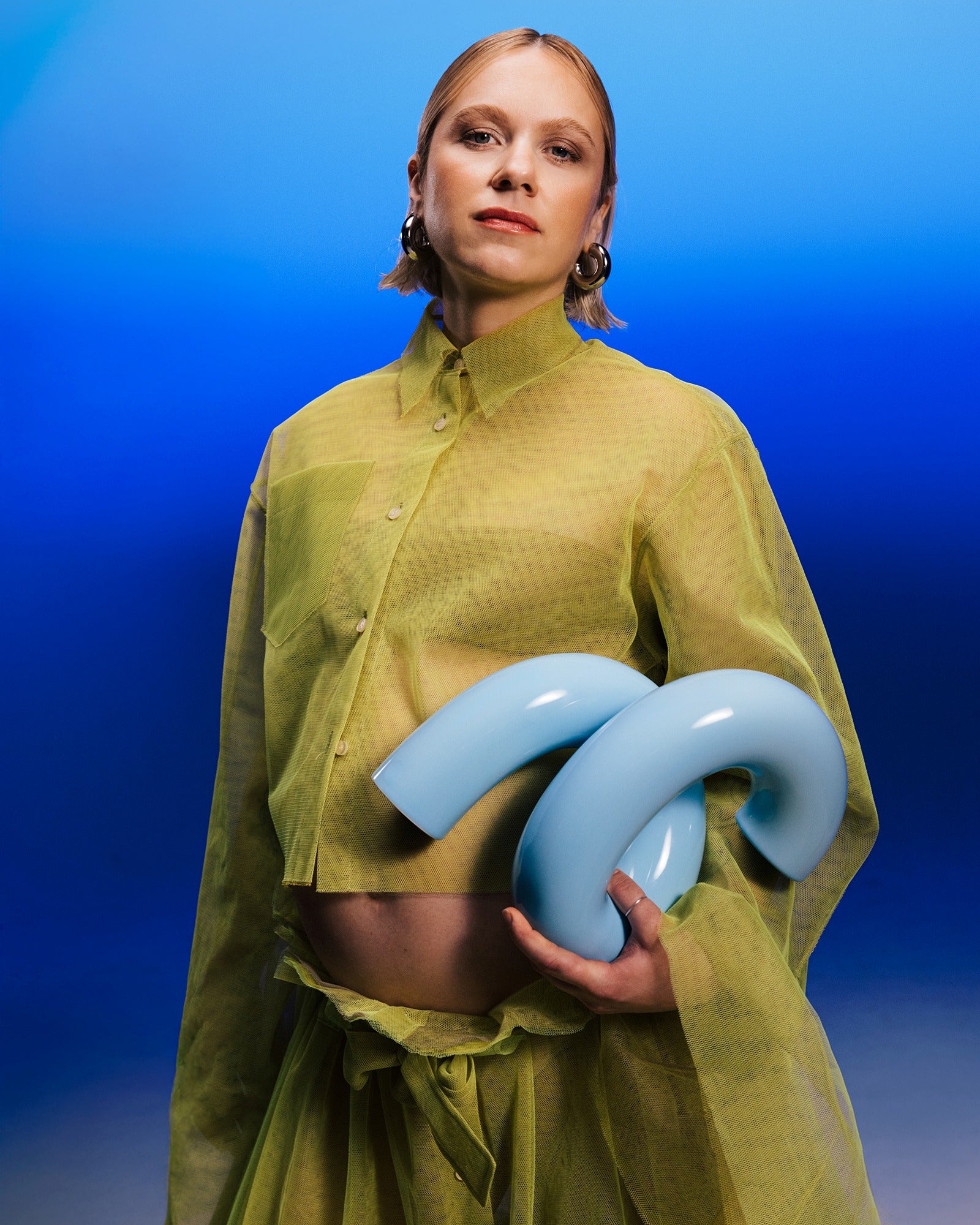 Frau in gelber Kleidung hält blaue, gebogene Objekte vor blauem Hintergrund.