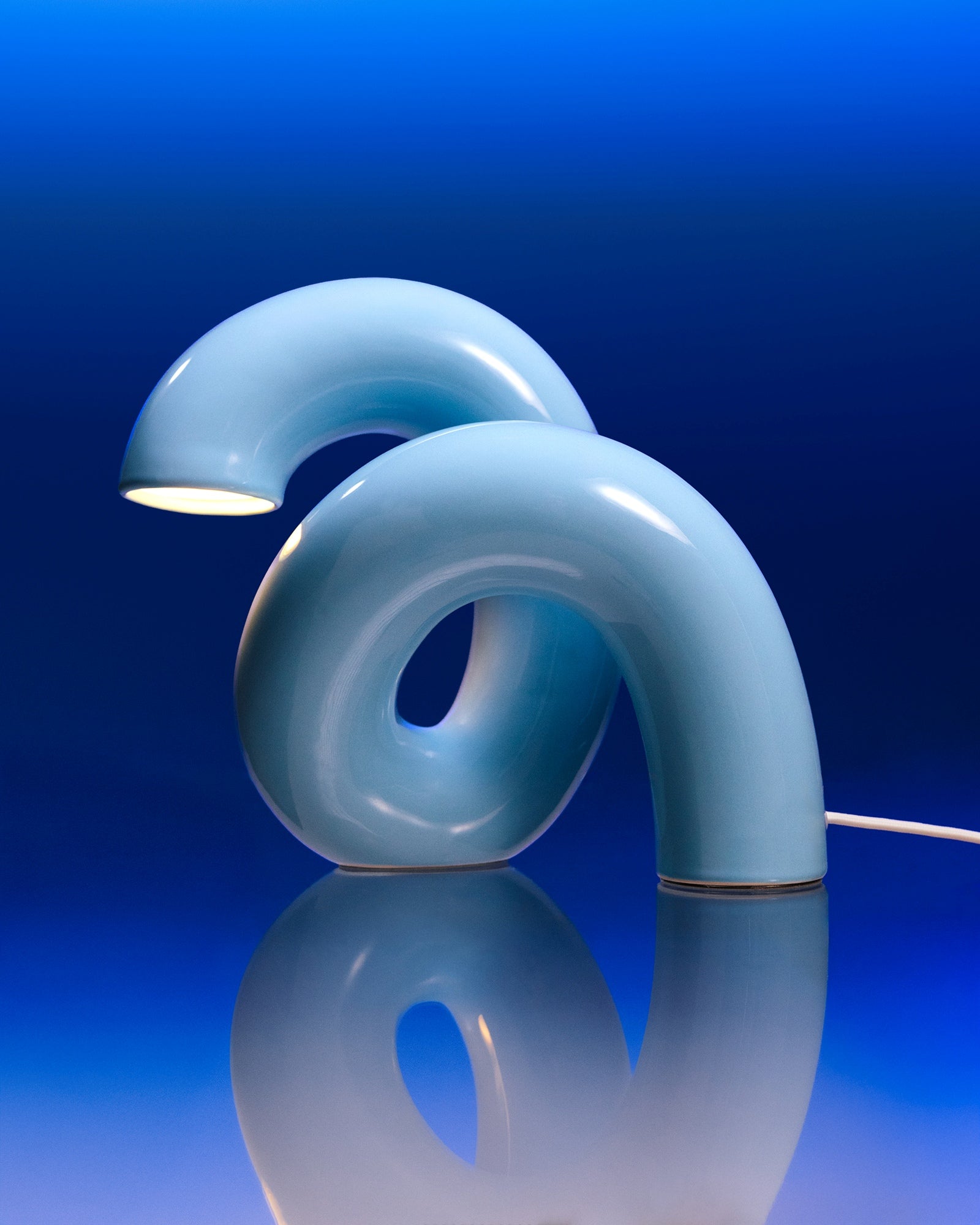 Moderne gebogene Tischlampe in Blau auf spiegelndem Untergrund vor blauem Hintergrund.