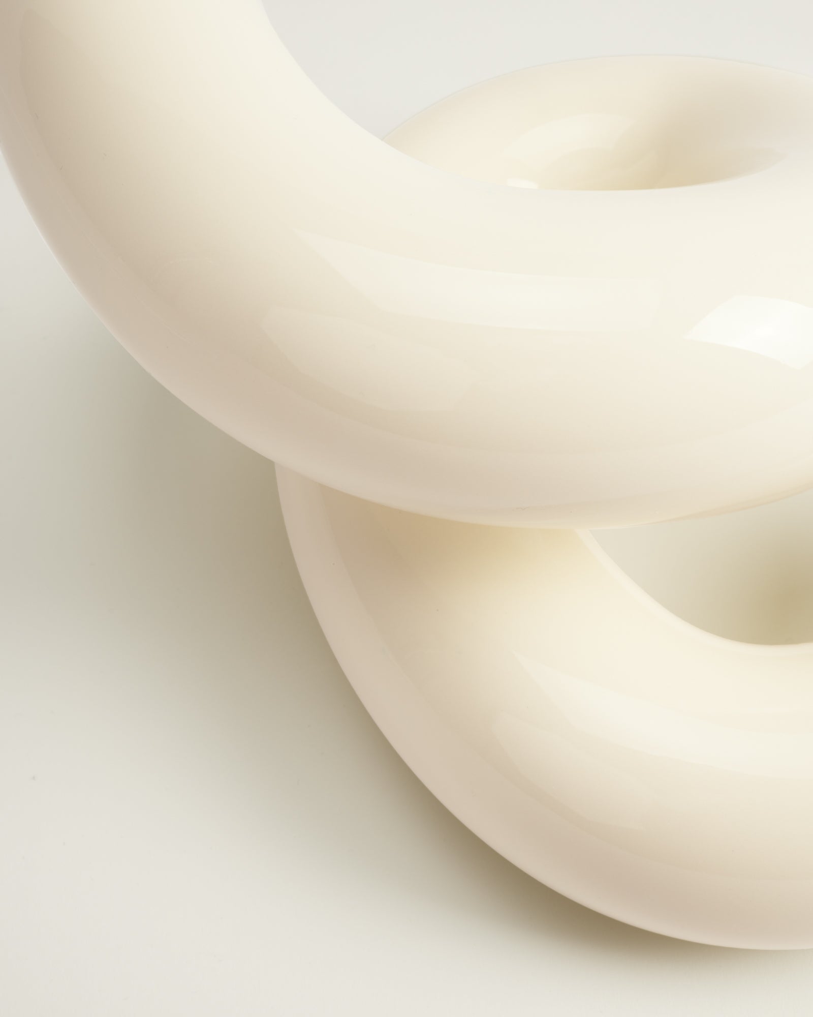 Zwei glänzende, cremefarbene Donut-förmige Objekte, die ineinander verschlungen sind, auf hellem Hintergrund.