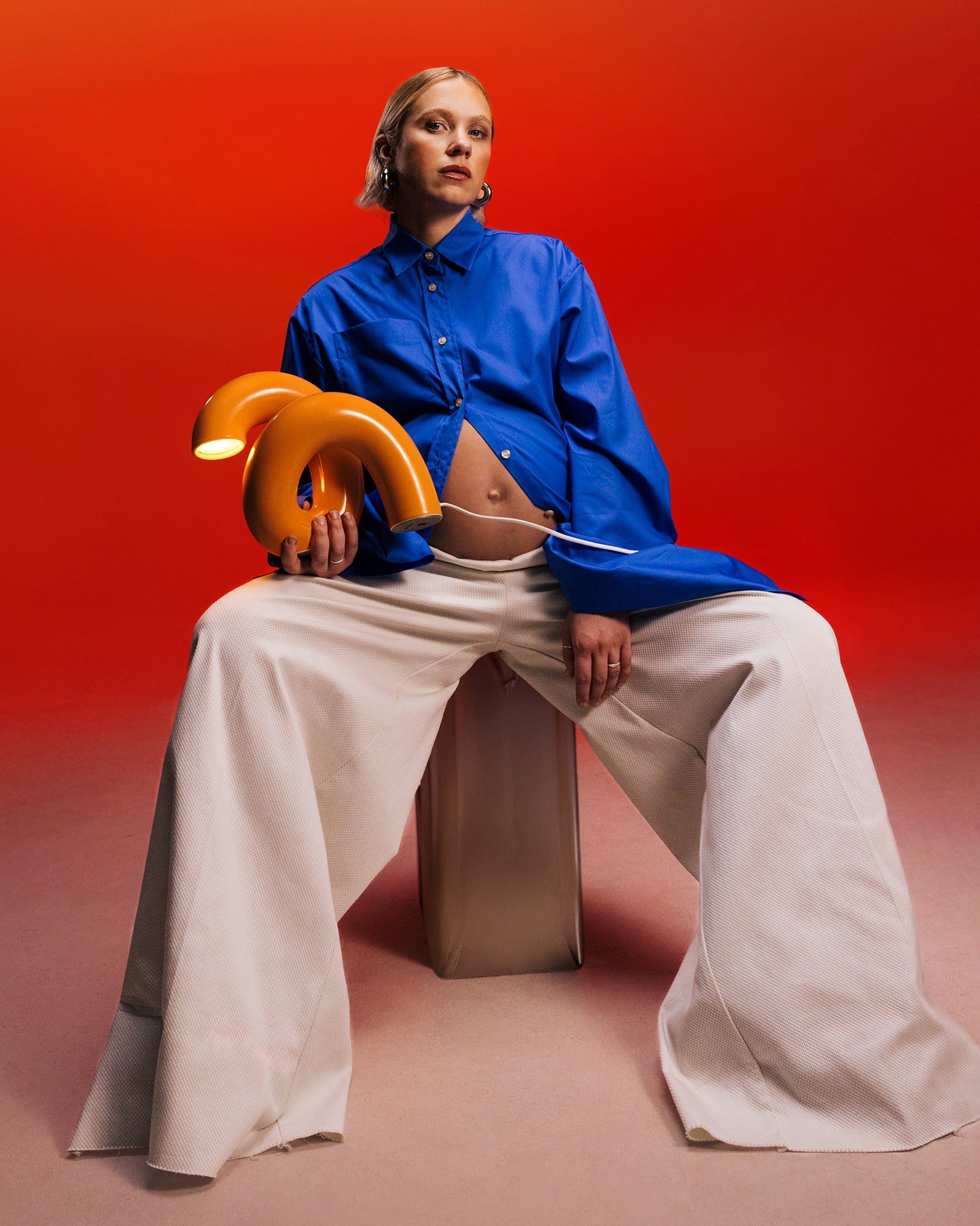 Person in blauer Bluse und weißen weiten Hosen sitzt auf Hocker, hält oranges Objekt, vor rotem Hintergrund.