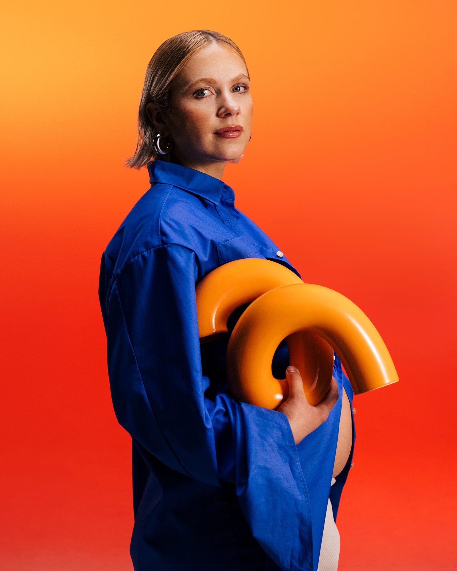 Frau in blauem Hemd hält ein gelbes, gebogenes Objekt vor einem orange-roten Hintergrund.