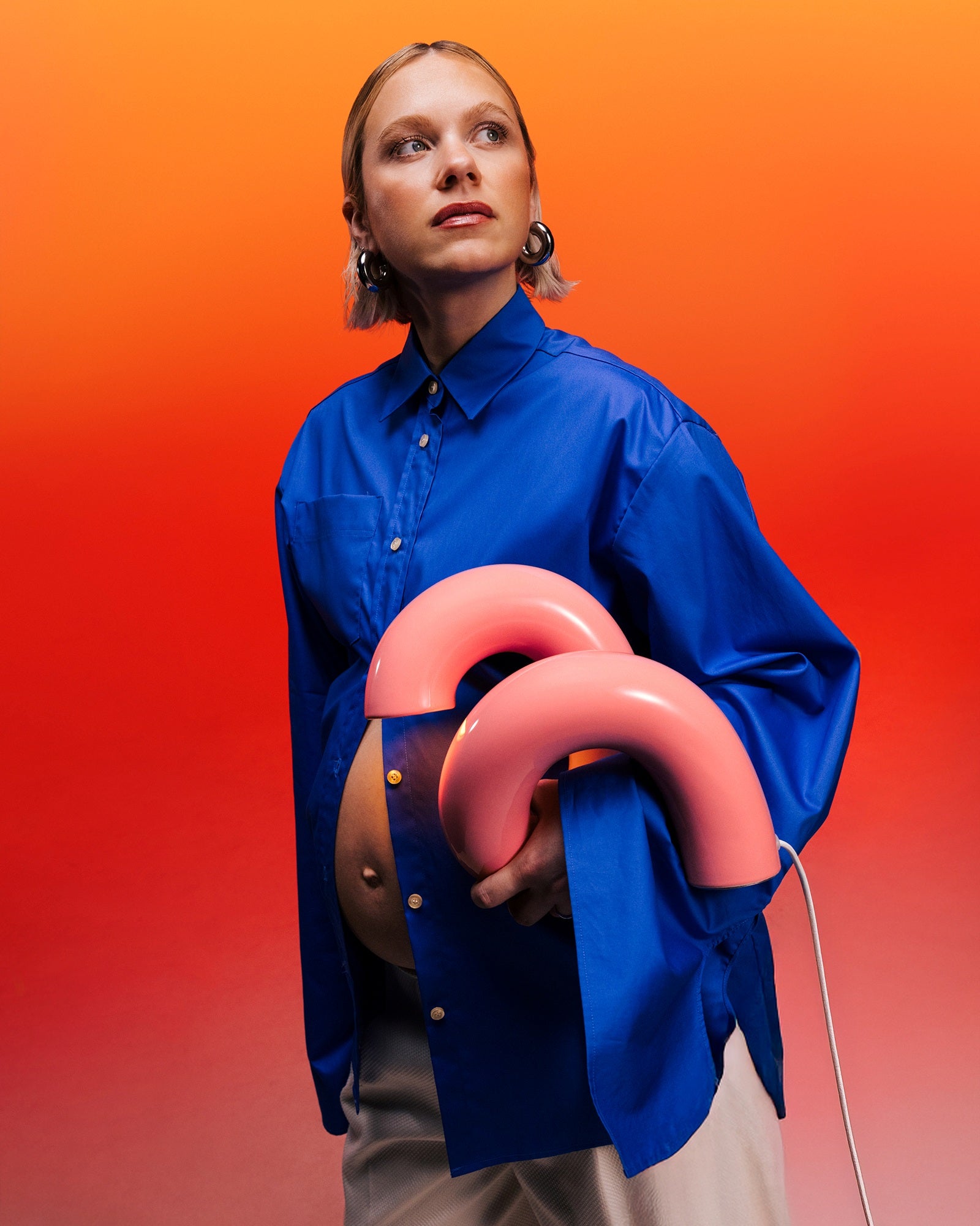 Schwangere Frau in blauem Hemd hält zwei rosa Objekte vor einem orangefarbenen Hintergrund.