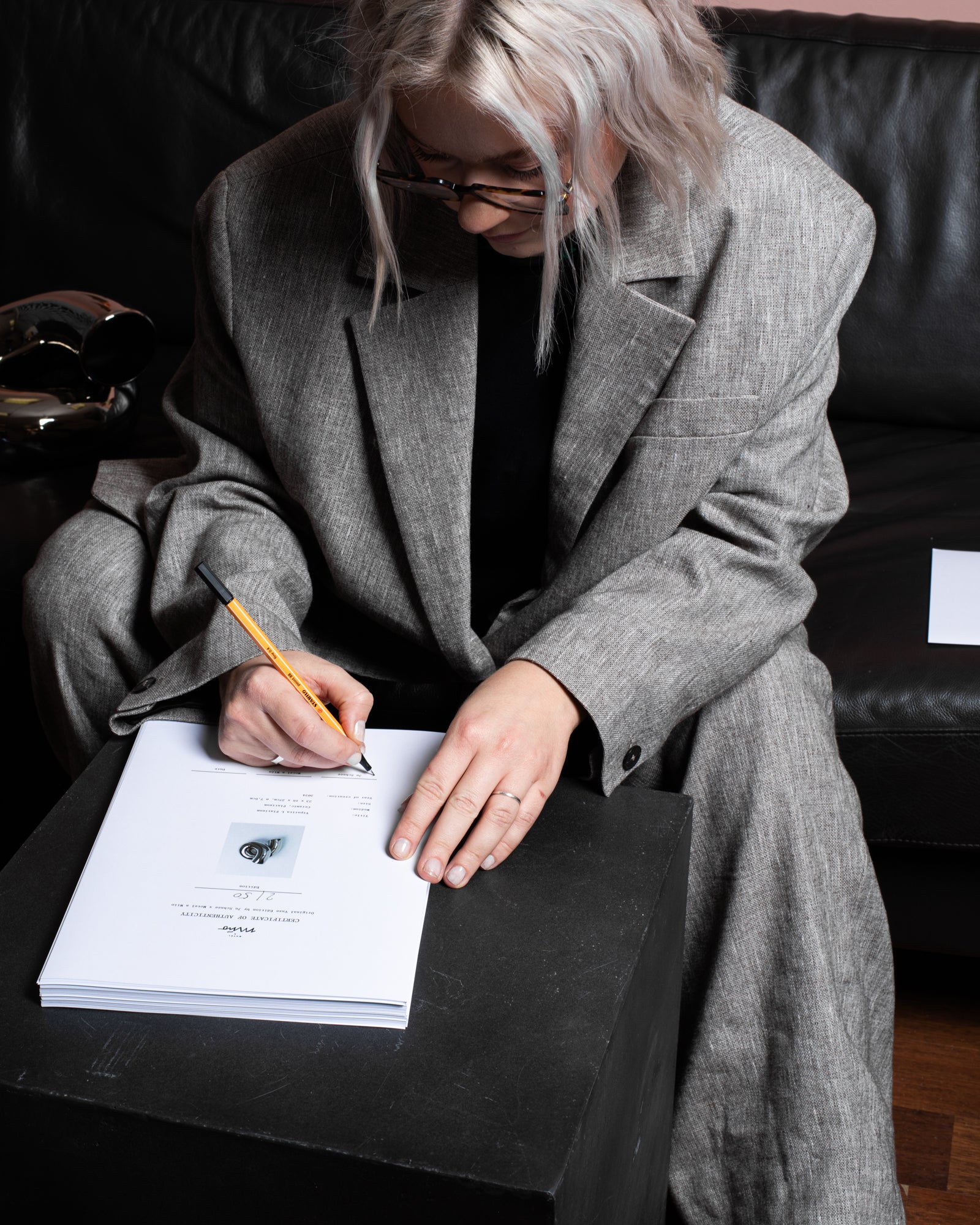 Person in grauem Anzug signiert ein Dokument auf schwarzem Tisch, mit einer Skulptur im Hintergrund.