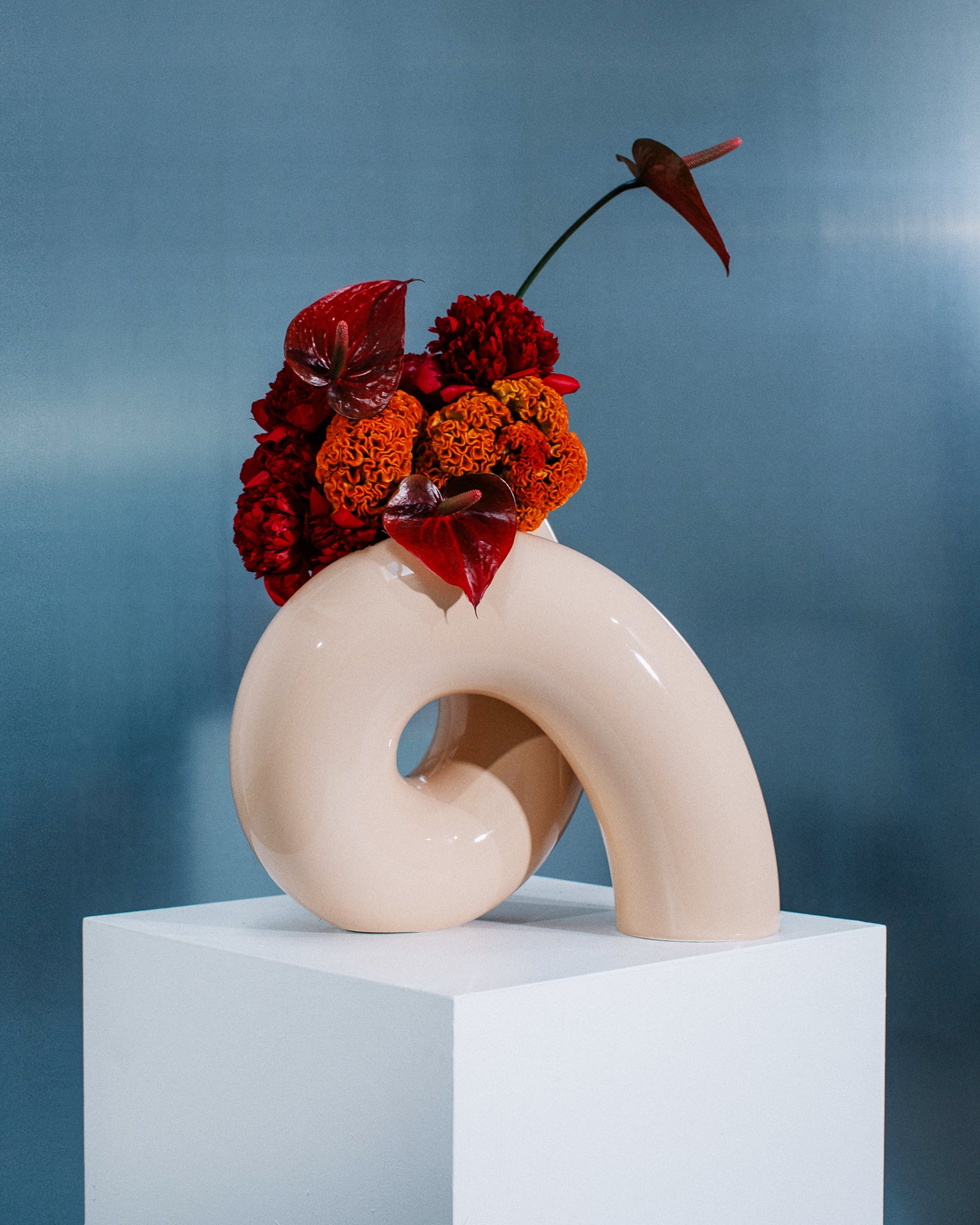 Abstrakte Vase auf Sockel mit roten und orangefarbenen Blumen, vor blauem Hintergrund.