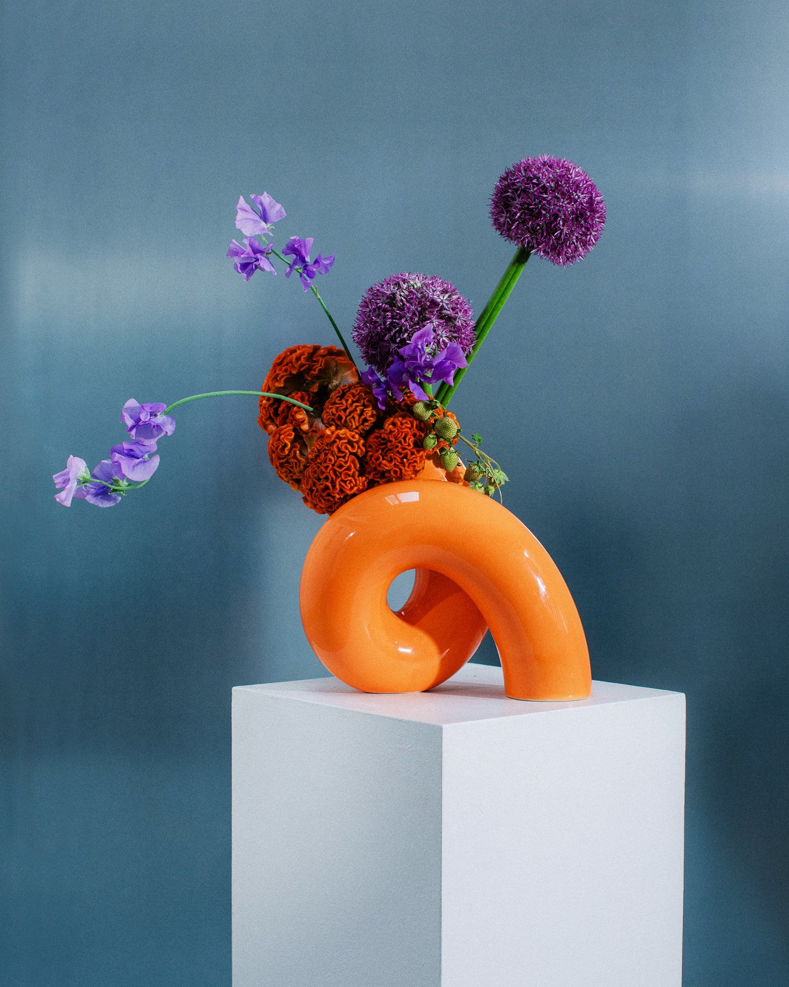 Orangene, geschwungene Vase auf Sockel mit lila und orangefarbenen Blumen vor blauem Hintergrund.
