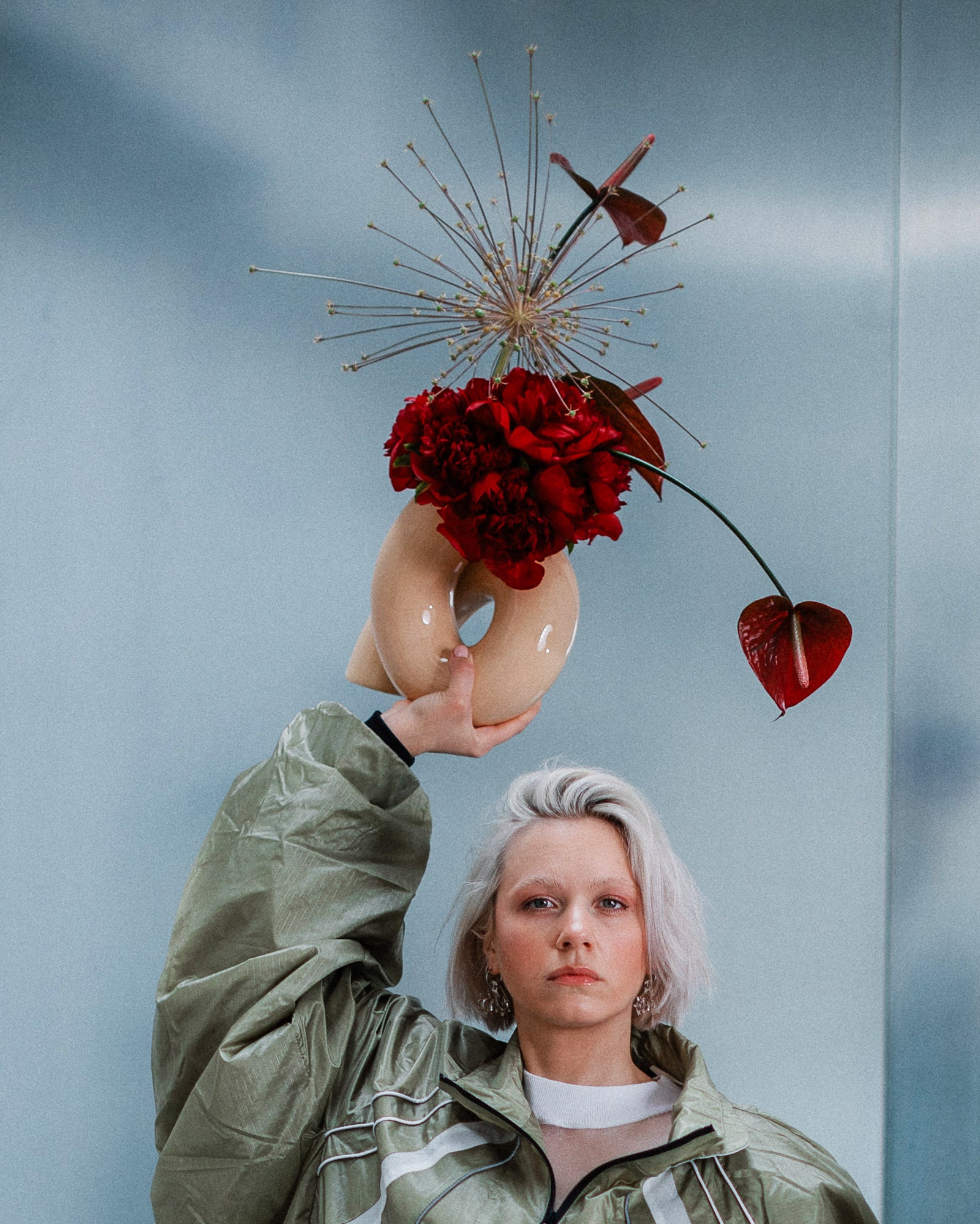 Eine Person in grüner Jacke hält eine Vase mit roten Blumen und geometrischem Design vor einem grauen Hintergrund.
