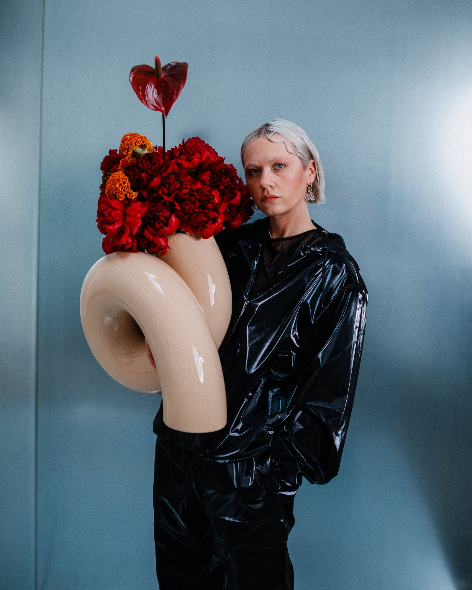 Person in schwarzem PVC-Anzug hält kunstvolle Vase mit roten Blumen und gebogenem, cremefarbenem Rohr.