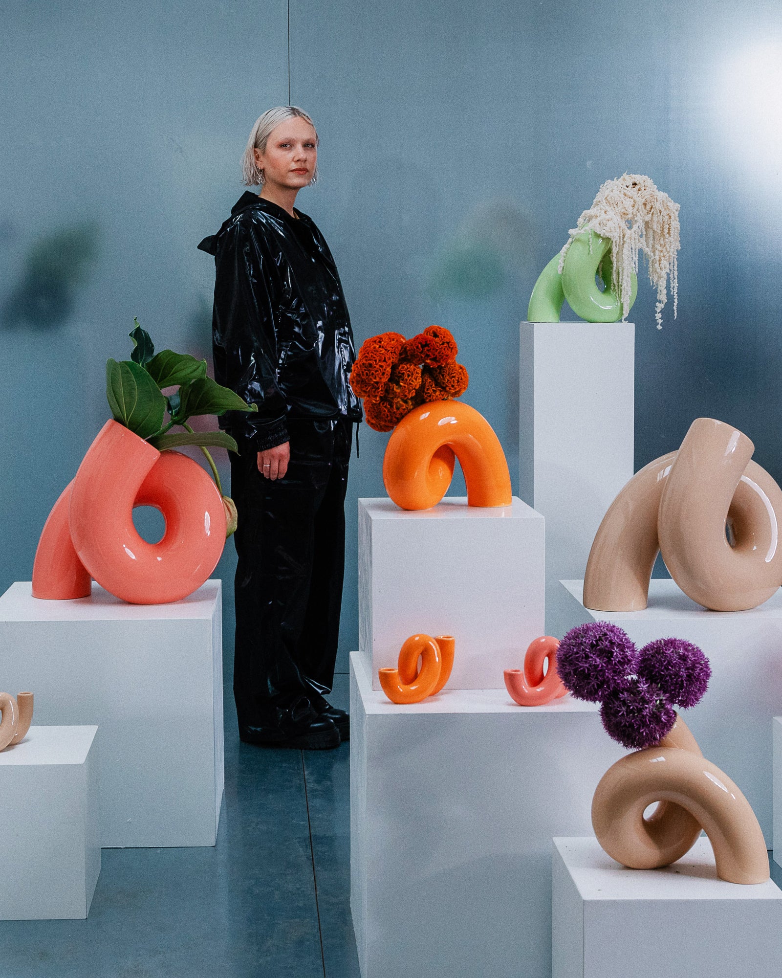 Eine Person steht neben abstrakten Skulpturen in verschiedenen Farben und Formen auf weißen Sockeln.