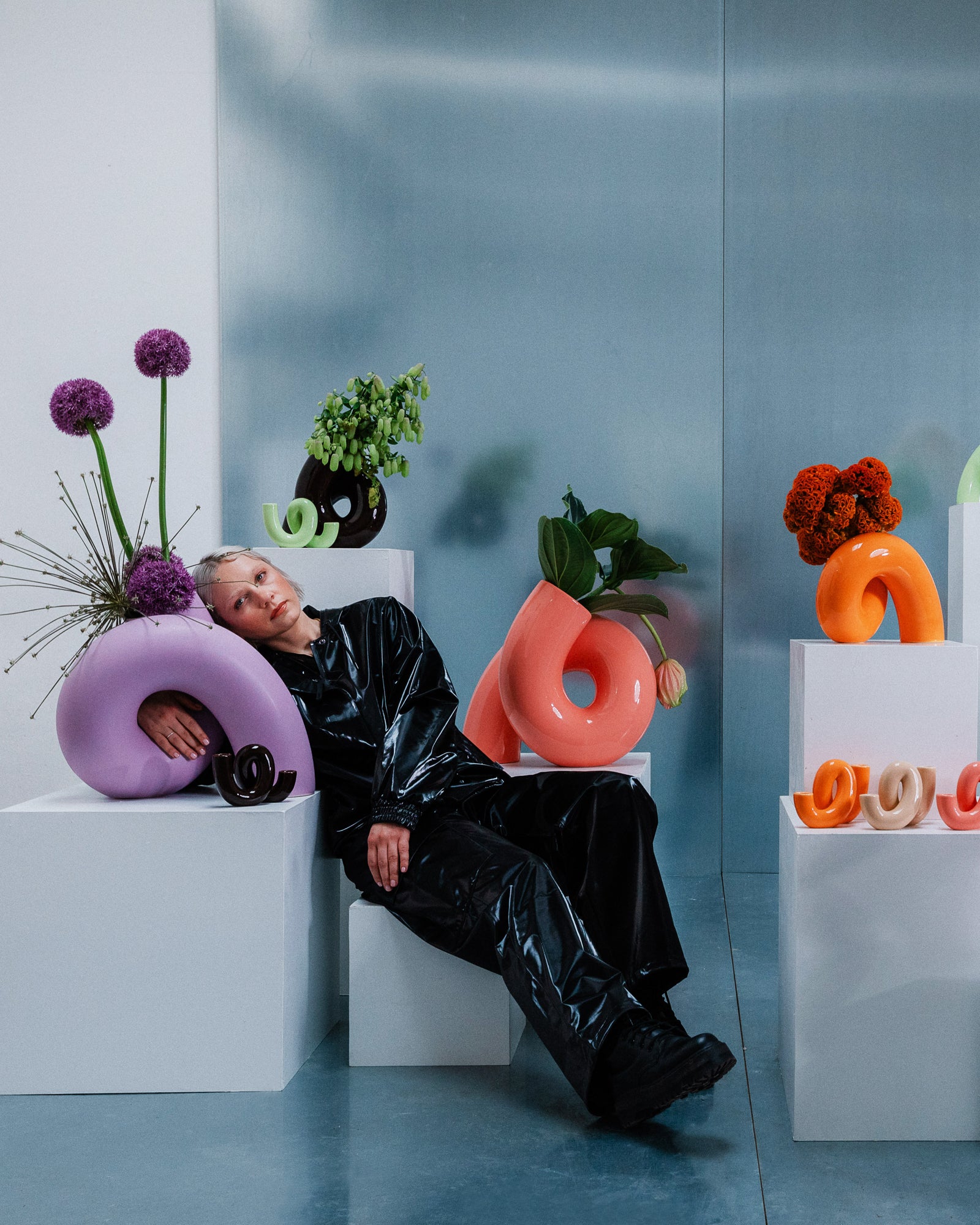 Eine Person in schwarzer Kleidung lehnt sich an skulpturale Blumenarrangements auf weißen Podesten.