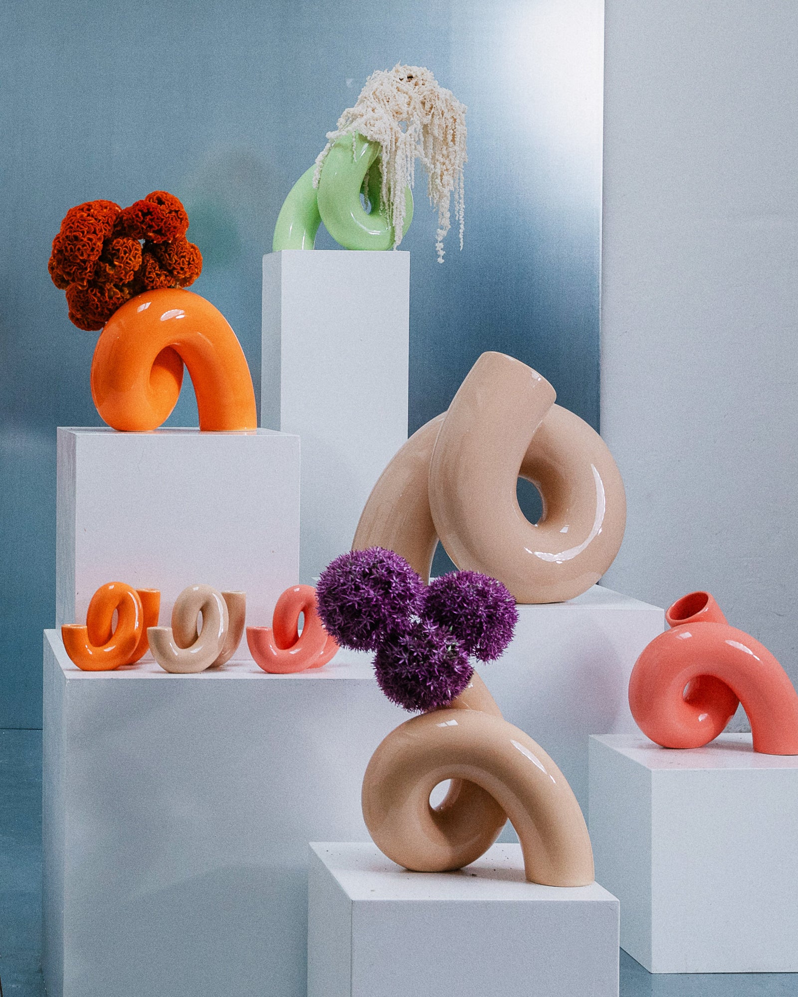 Verschiedene bunte, schlaufenartige Keramikskulpturen auf weißen Sockeln, dekoriert mit Blumen in einem modernen Ausstellungsraum.