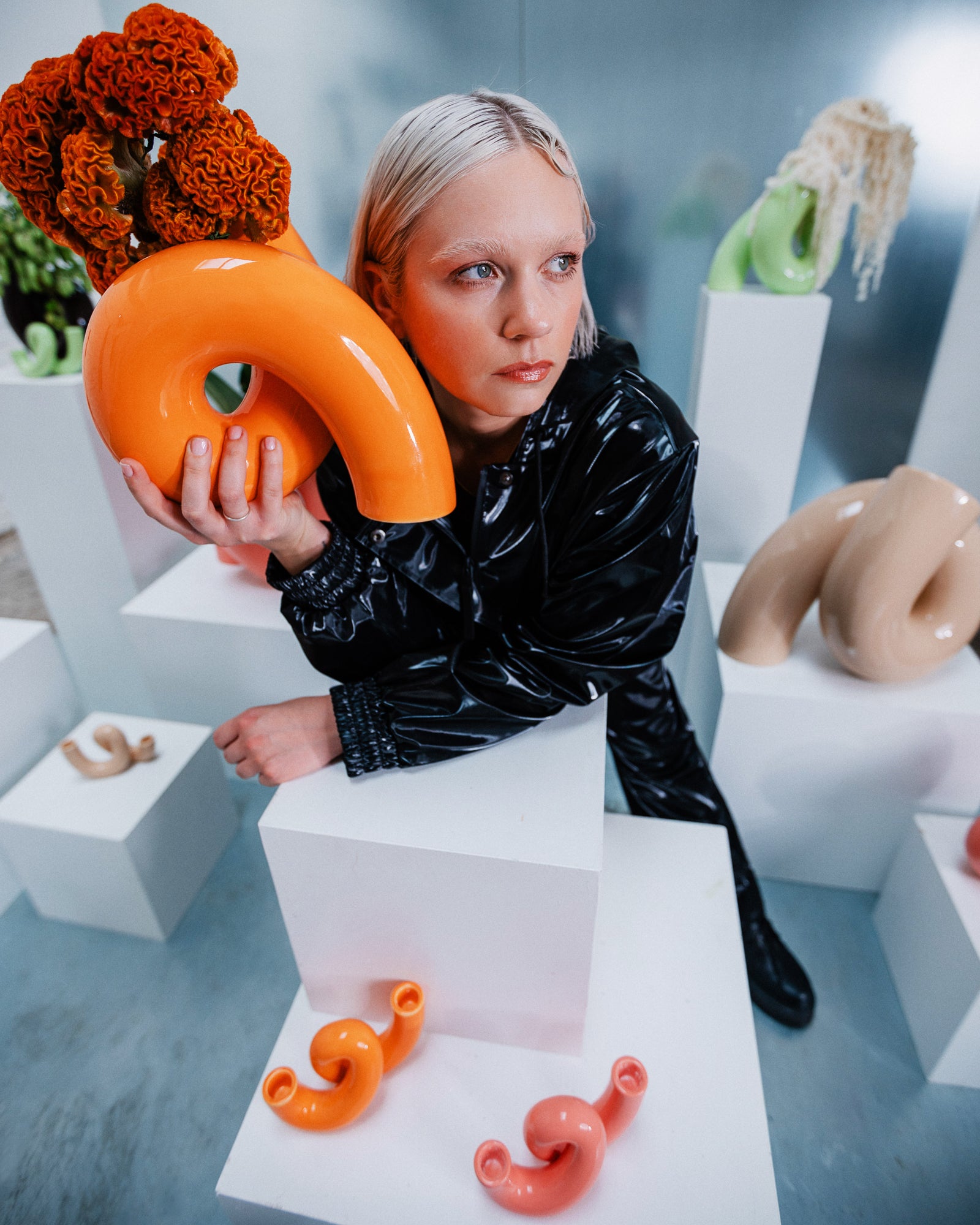 Eine Person in schwarzer Kleidung hält eine orangefarbene Skulptur und sitzt zwischen abstrakten Kunstwerken auf weißen Podesten.