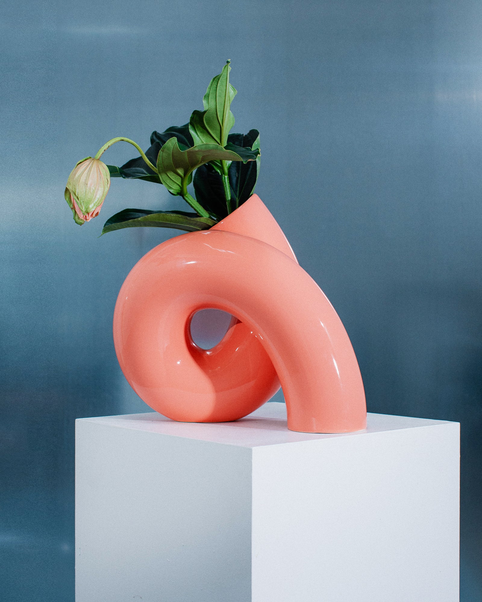 Rosa, geschwungene Vase auf Sockel mit grünen Blättern und einer geschlossenen Blüte, vor grauem Hintergrund.