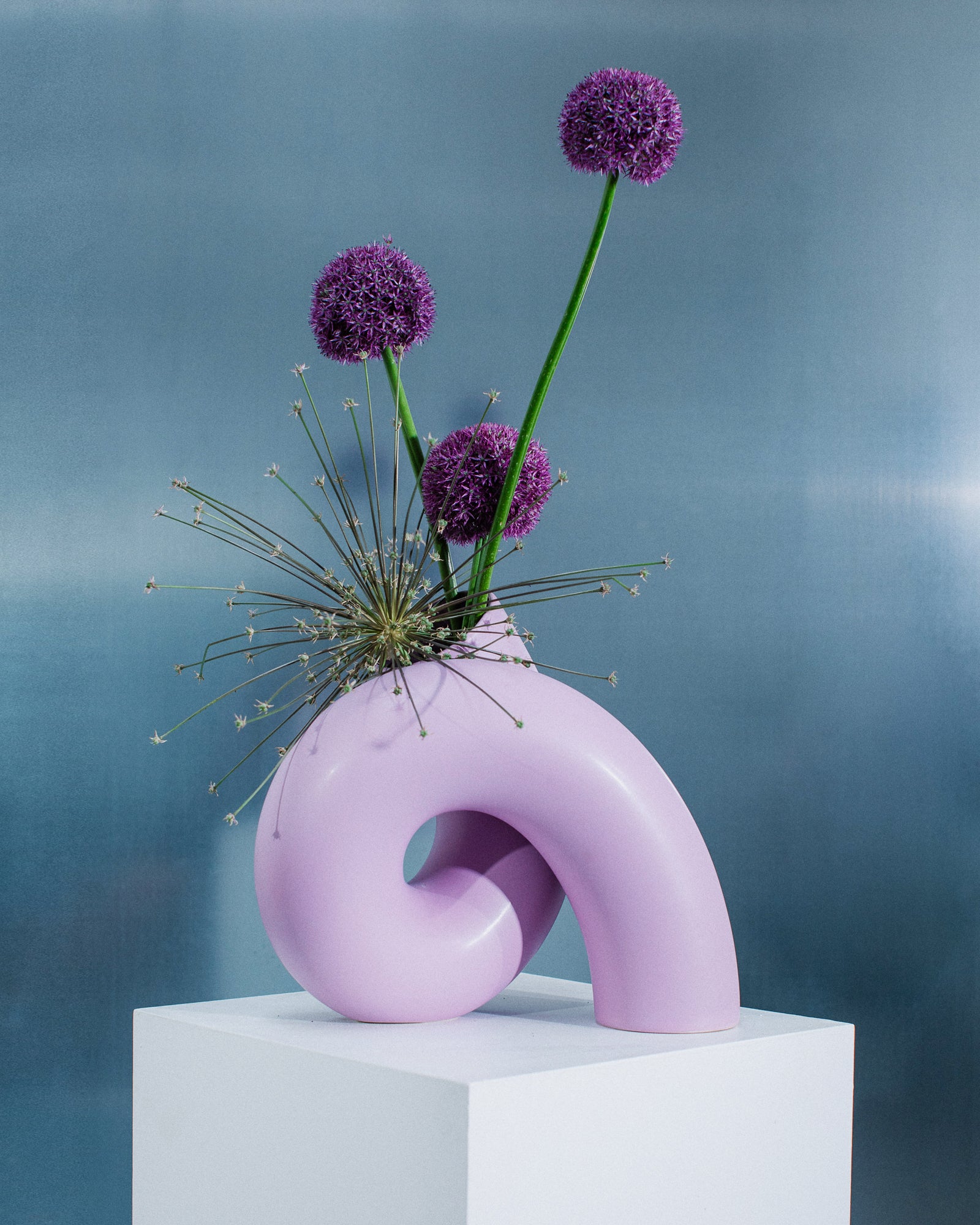 Eine modern geformte, lilafarbene Vase mit violetten Kugelblumen steht auf einem weißen Podest vor grauem Hintergrund.