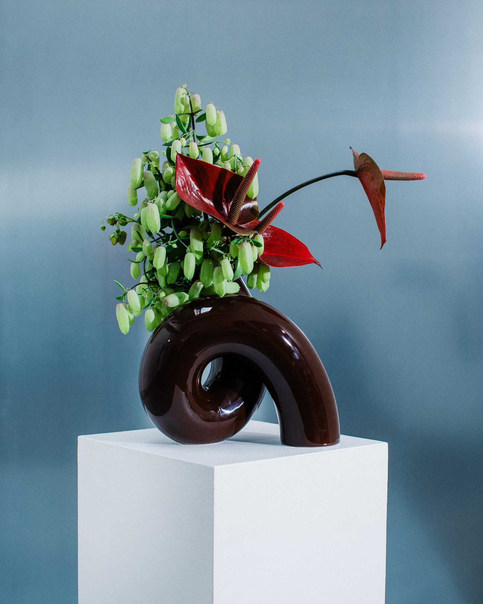 Moderne Skulpturvase mit grünen und roten Blumen auf einem weißen Sockel vor blauem Hintergrund.