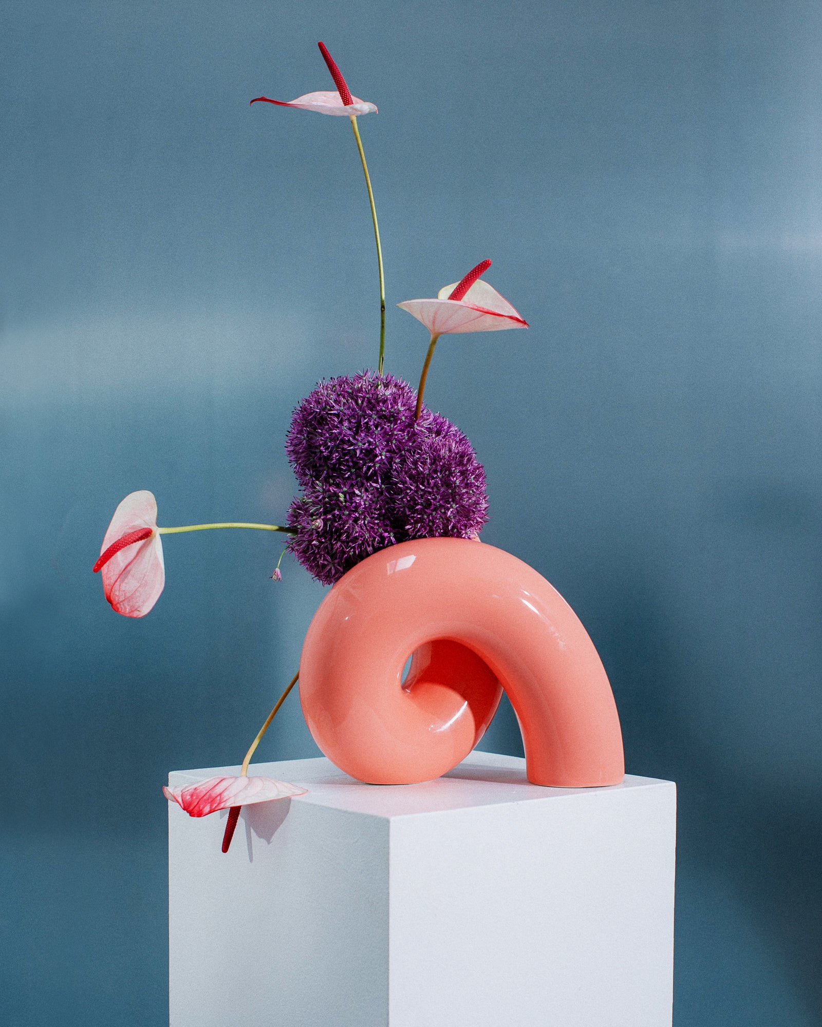 Rote Keramikskulptur und violette Blüten auf weißem Podest vor blauem Hintergrund.