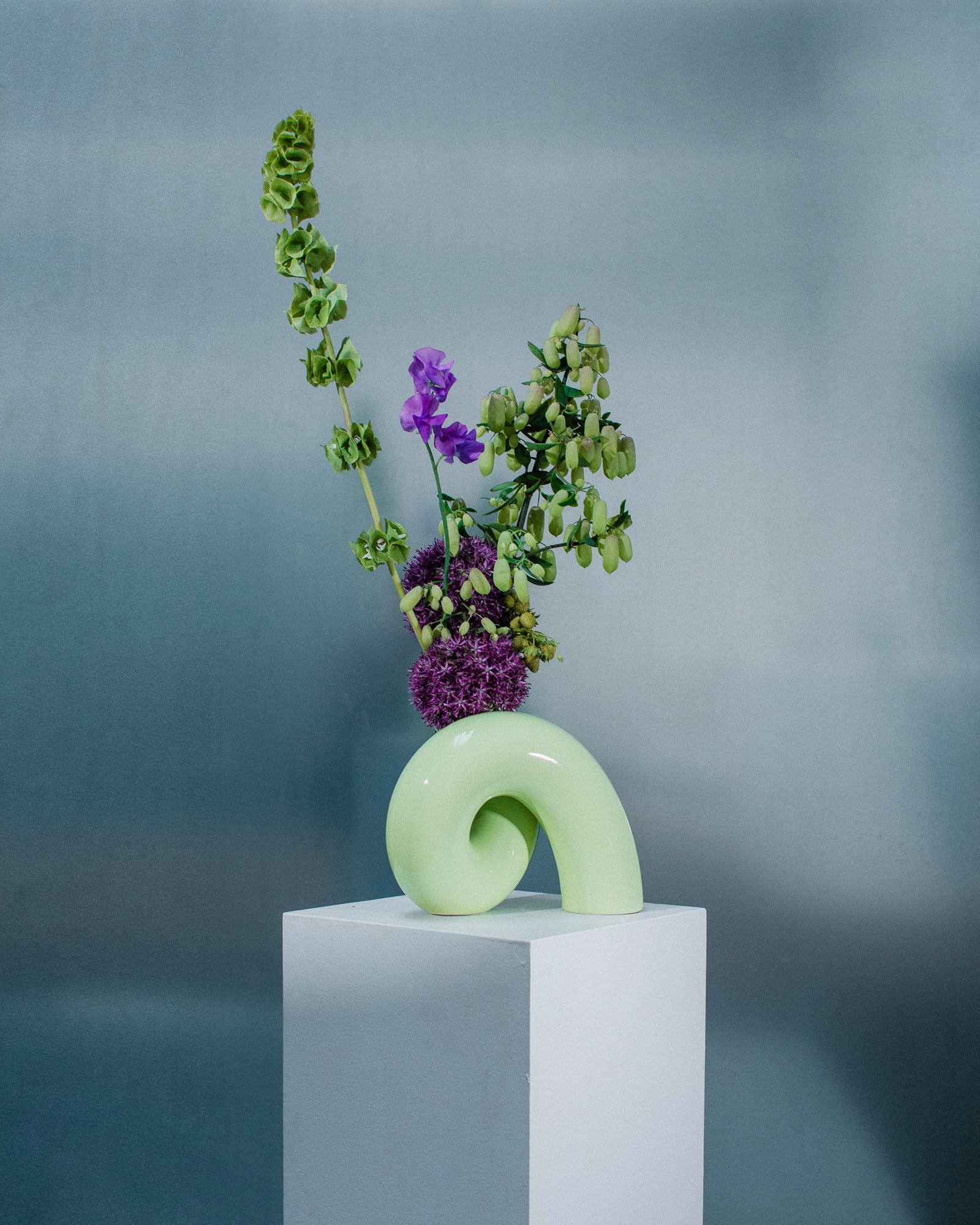 Grüne, geschwungene Vase mit langen grünen und violetten Blumen auf weißem Podest vor grauem Hintergrund.