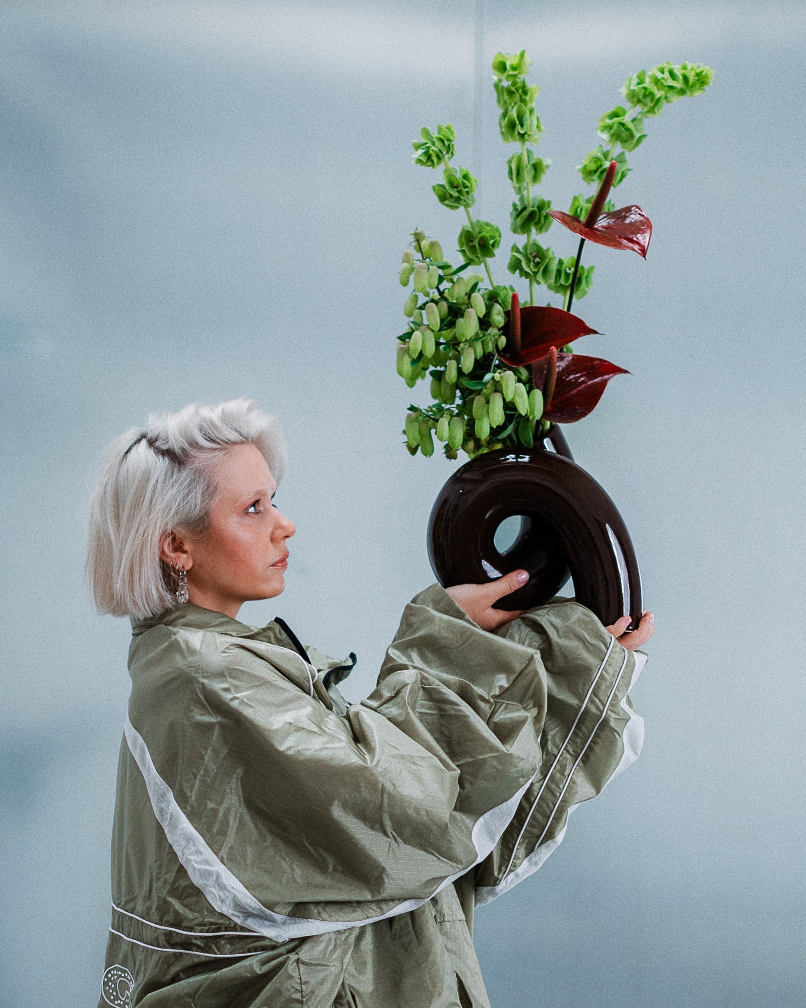 Frau mit weißem Haar hält eine schwarze Vase mit grünen und roten Blumen vor einem blaugrauen Hintergrund.