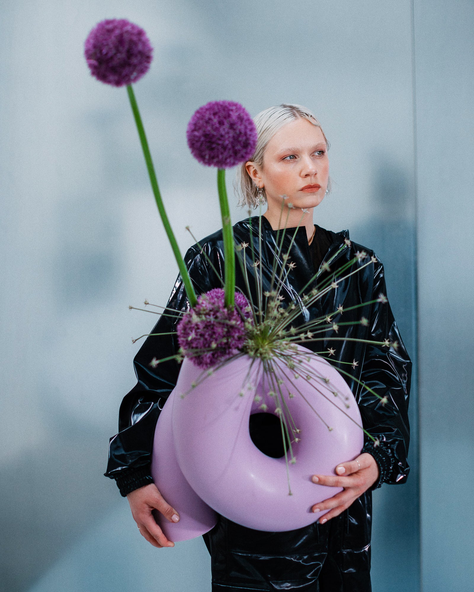 Person in schwarzem Outfit hält eine große, lilafarbene Skulptur mit langen Stielblumen.