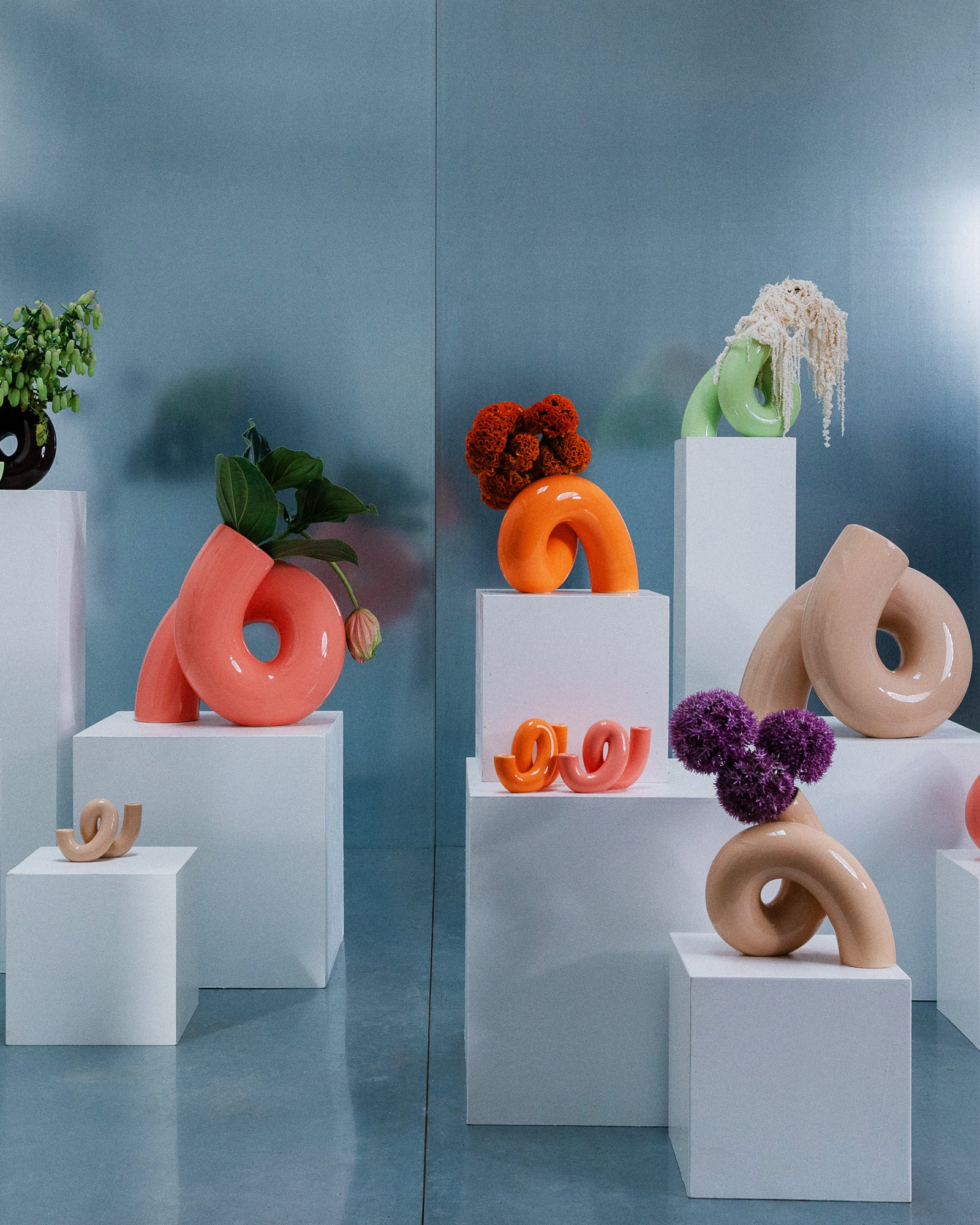 Farbige, skulpturale Vasen in unterschiedlichen Formen und Größen mit Blumen, auf verschieden hohen weißen Sockeln.