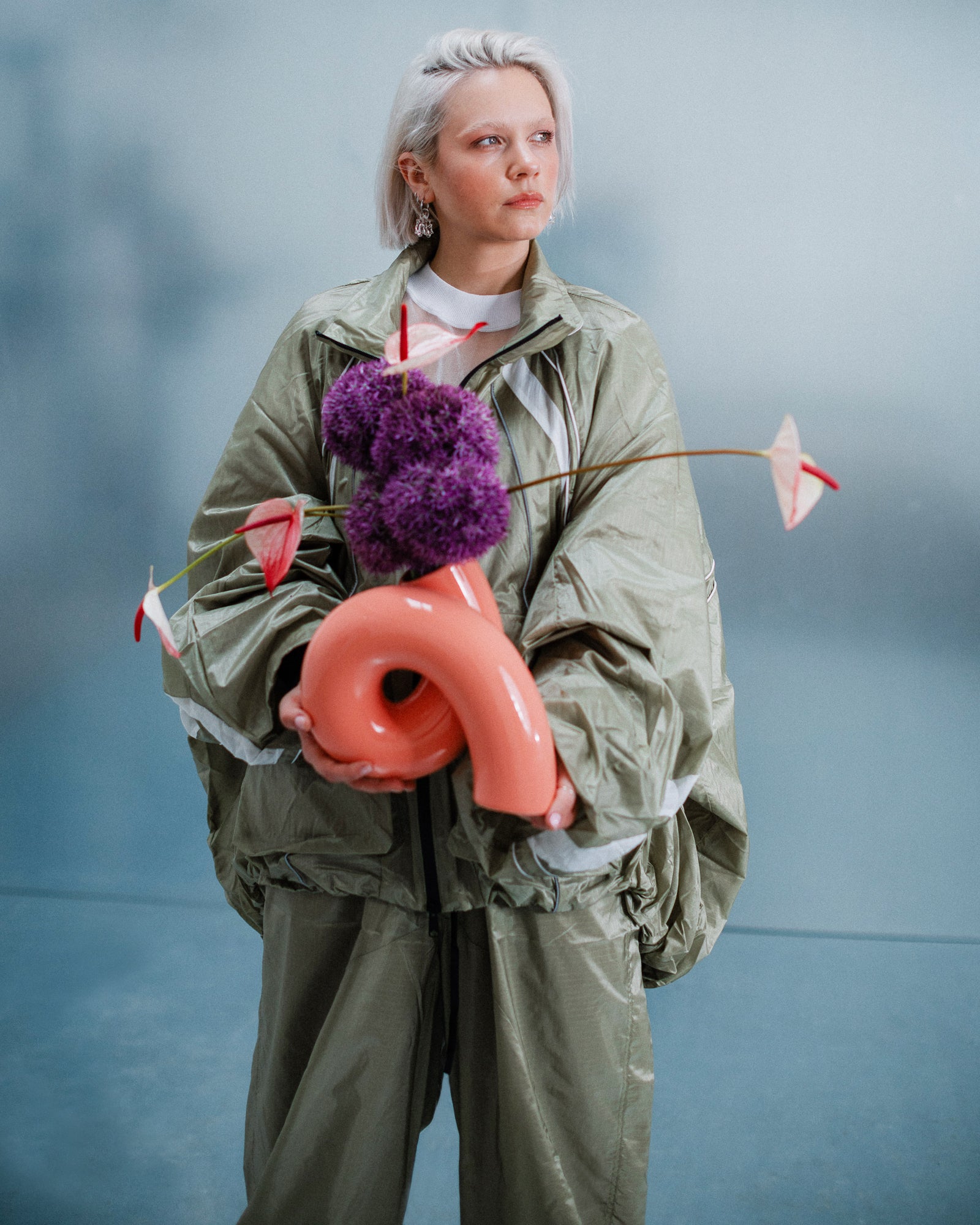 Person in grüner Kleidung hält eine korallenfarbene Vase mit lila und rosa Blumen vor einem unscharfen Hintergrund.
