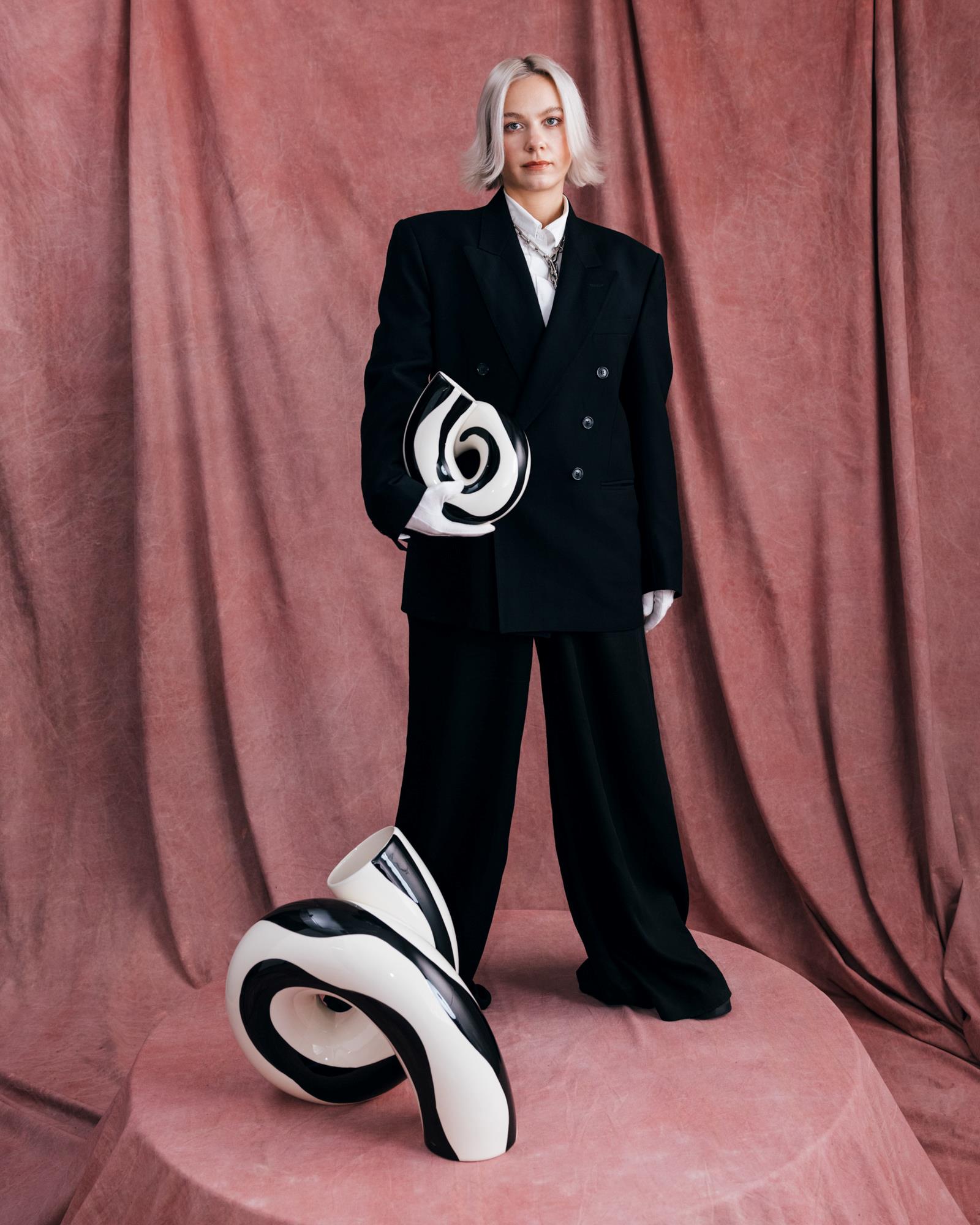 Person in einem schwarzen Anzug hält eine abstrakte, schwarz-weiße Skulptur auf einem rosa Podest mit rosafarbenem Hintergrund.