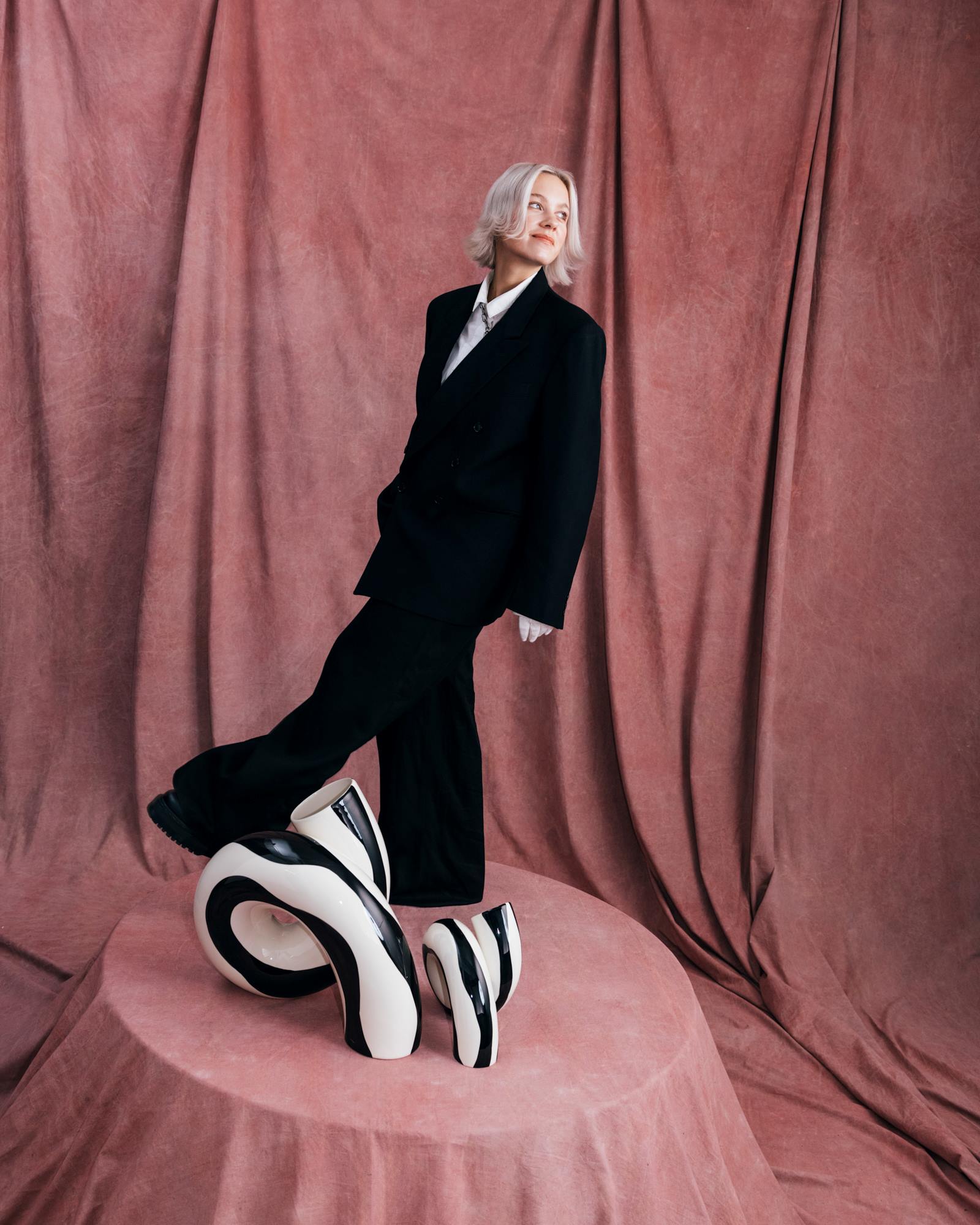 Eine Person in schwarzem Anzug steht lächelnd auf einem rosa Podest mit abstrakten Skulpturen, vor rosafarbenem Hintergrund.