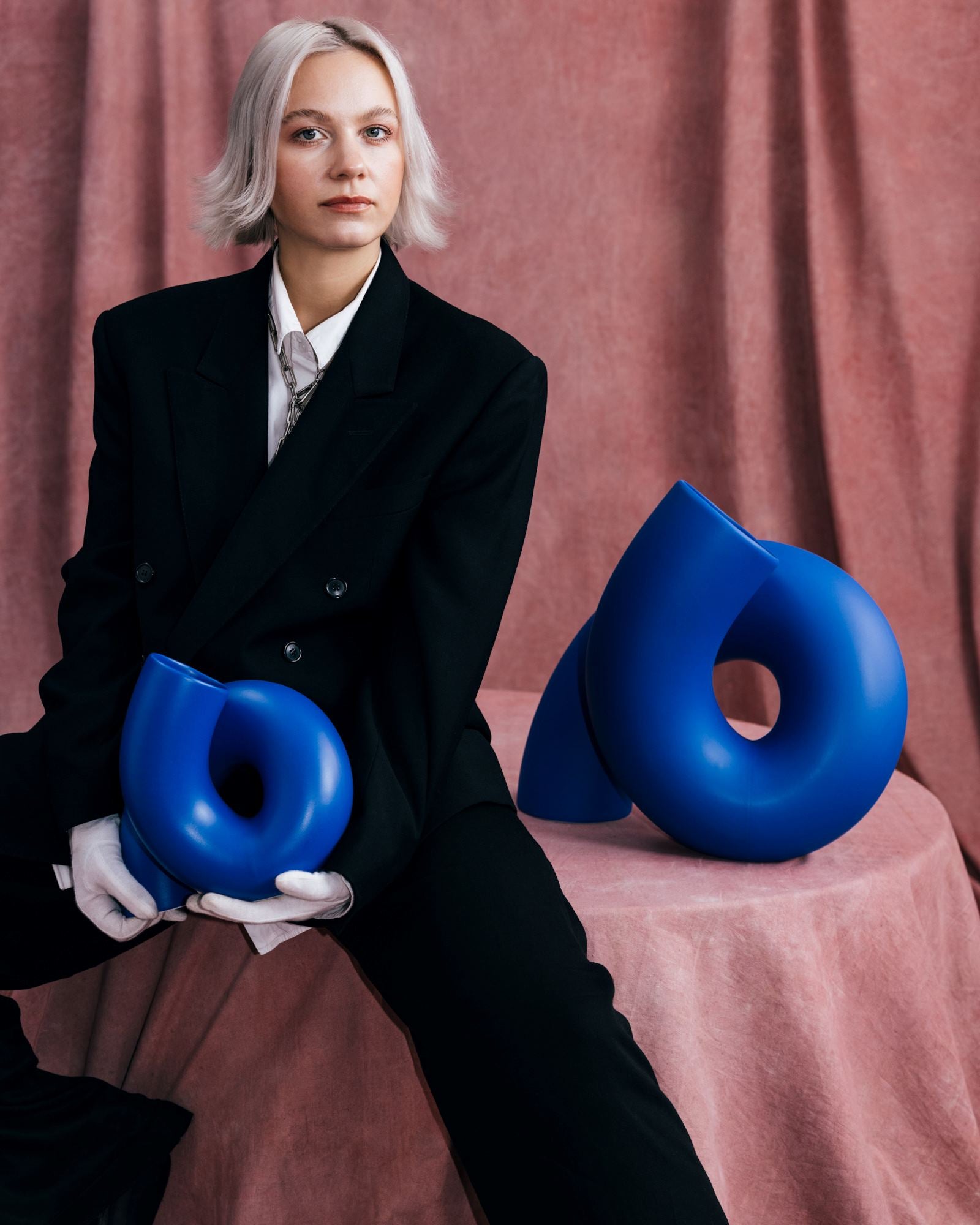 Person im schwarzen Anzug sitzt auf rosa Stoff, hält blaue abstrakte Skulptur, weitere Skulptur neben ihr.
