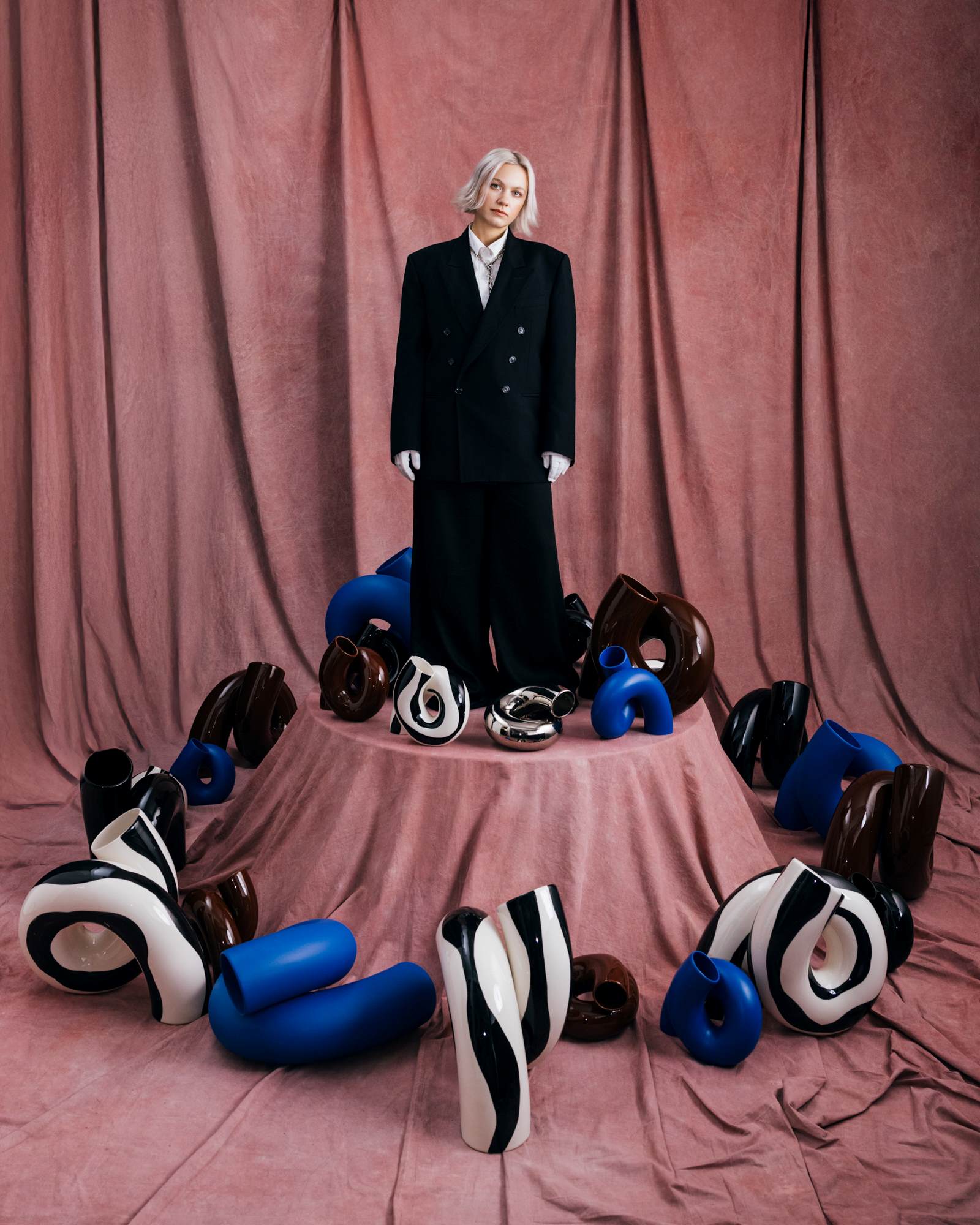 Person in schwarzem Anzug steht vor rosa Vorhang, umgeben von bunten, geschwungenen Keramikskulpturen.