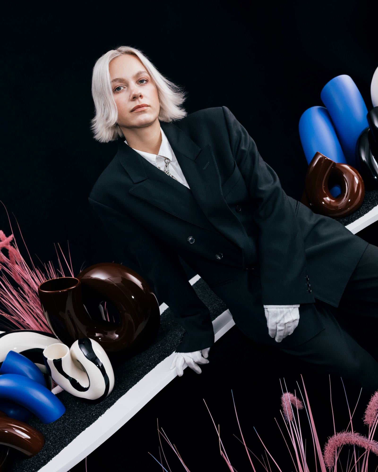 Person in schwarzem Anzug lehnt an einem Tisch mit modernen Vasen in Braun, Weiß und Blau, vor schwarzem Hintergrund.