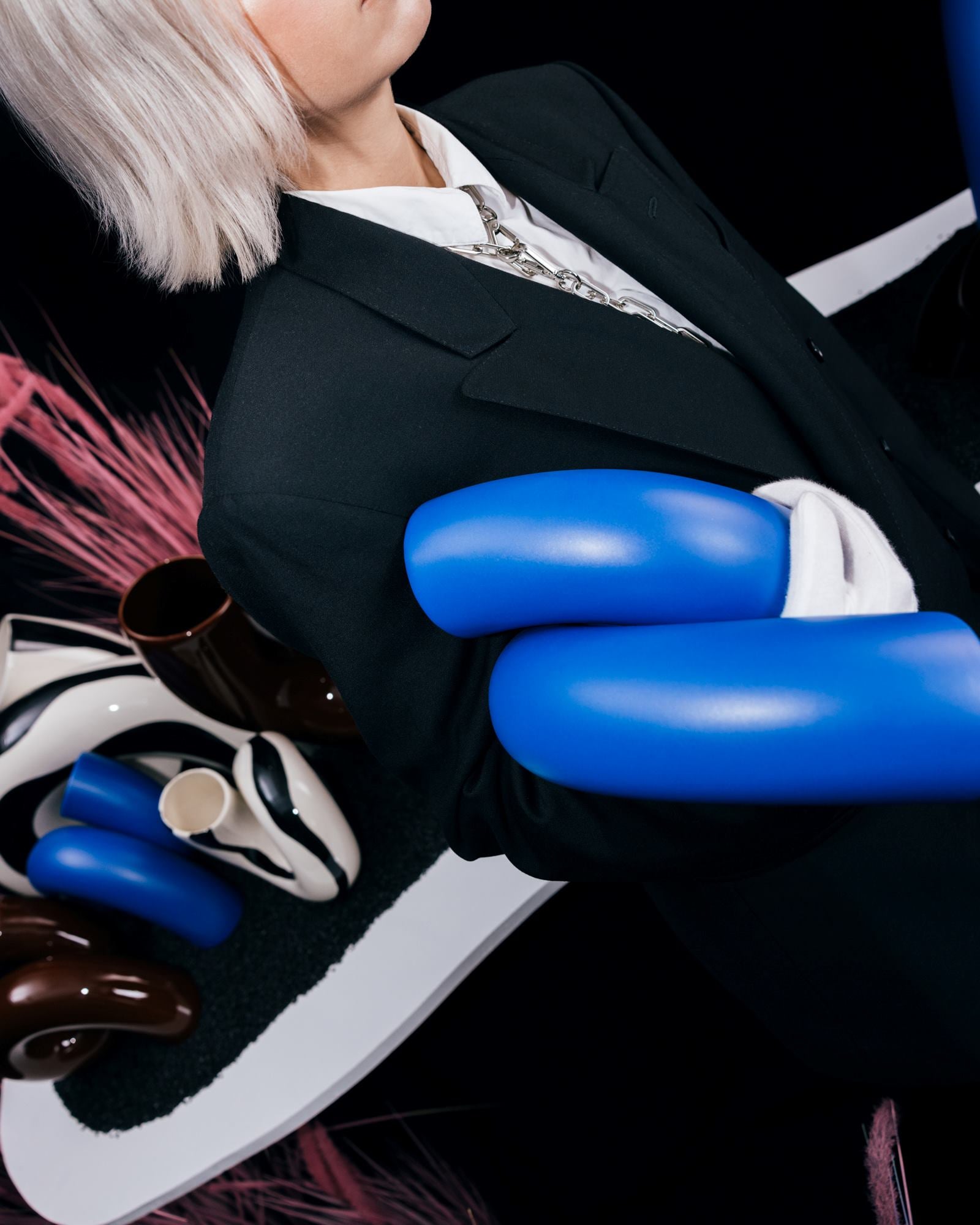 Person in schwarzem Anzug mit weißen und blauen Objekten vor einem abstrakten Hintergrund aus rosa und braunen Formen.