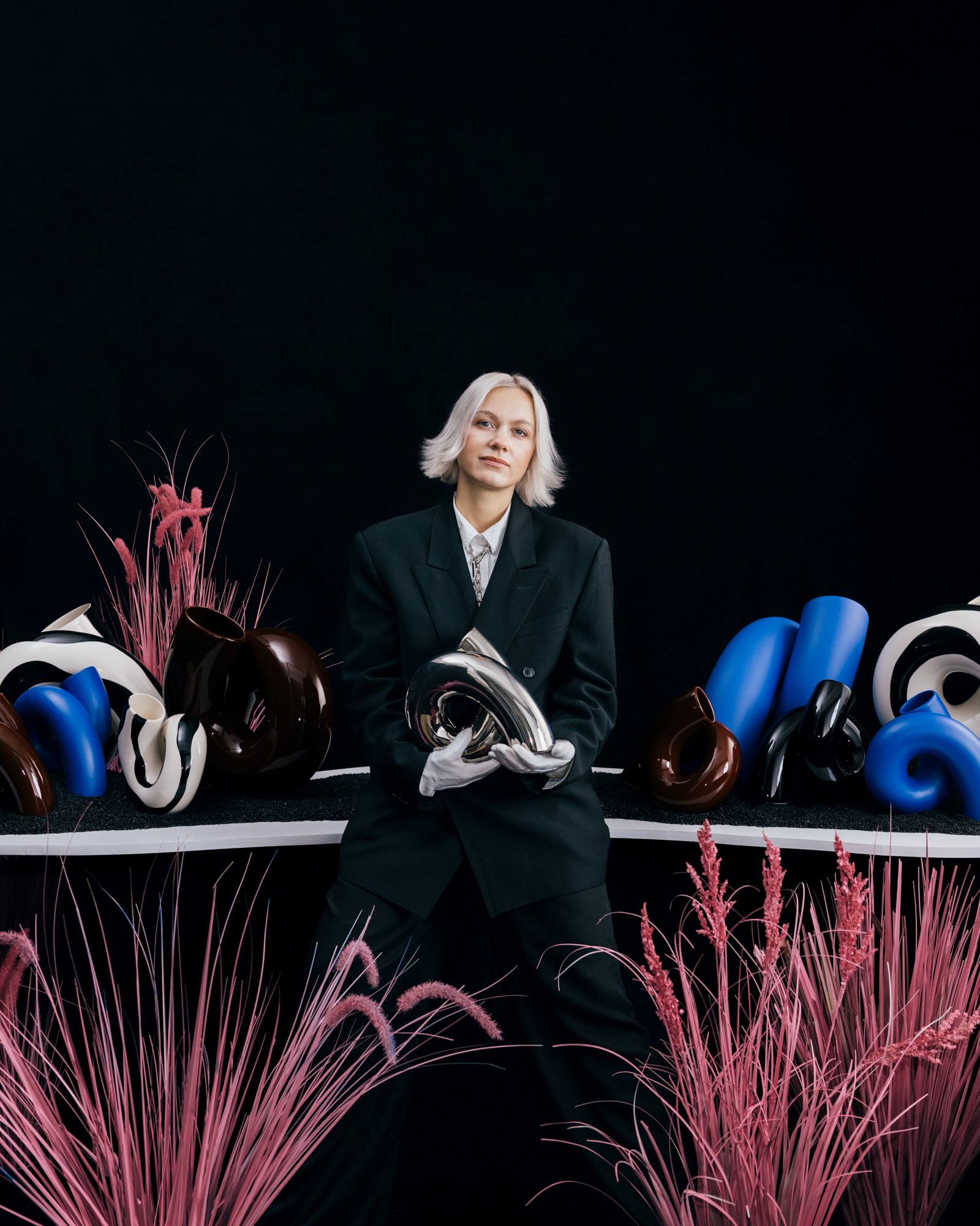 Person in schwarzem Anzug mit weißen Handschuhen hält Skulptur, umgeben von abstrakten Formen und rosa Pflanzen.